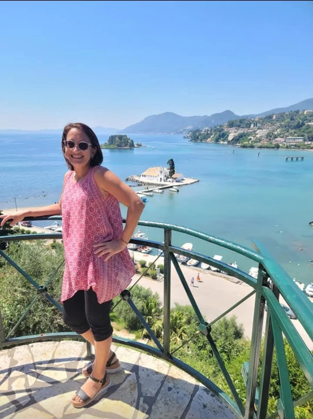 Woman in pink patterned sleeveless top and black capri pants standing on a viewing platform overlooking a large body of water with a small island, boats, and hills in the background under clear blue skies.