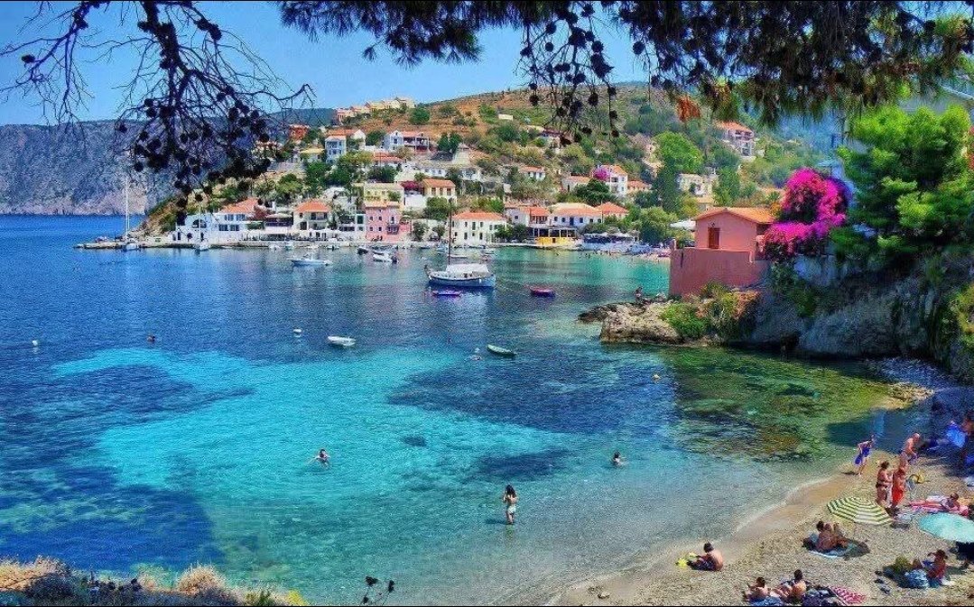 A lively beach scene with people swimming and relaxing near colorful houses on a hillside, over a bright blue ocean, with boats anchored and trees framing the view.