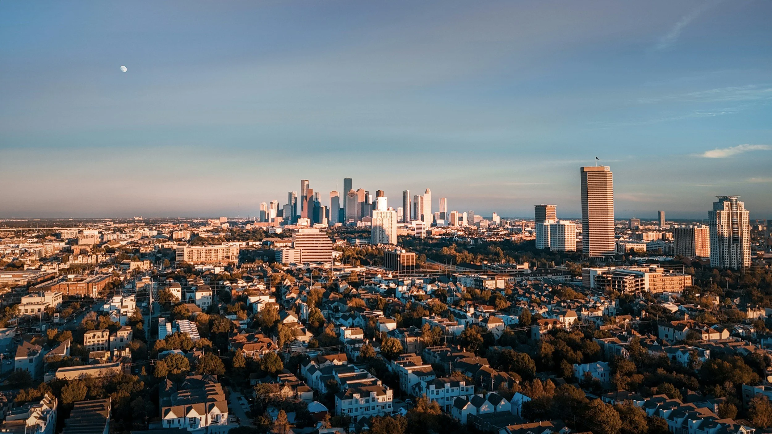A city skyline with tall buildings and a clear sky, with the moon visible in the top left corner, and residential houses in the foreground.