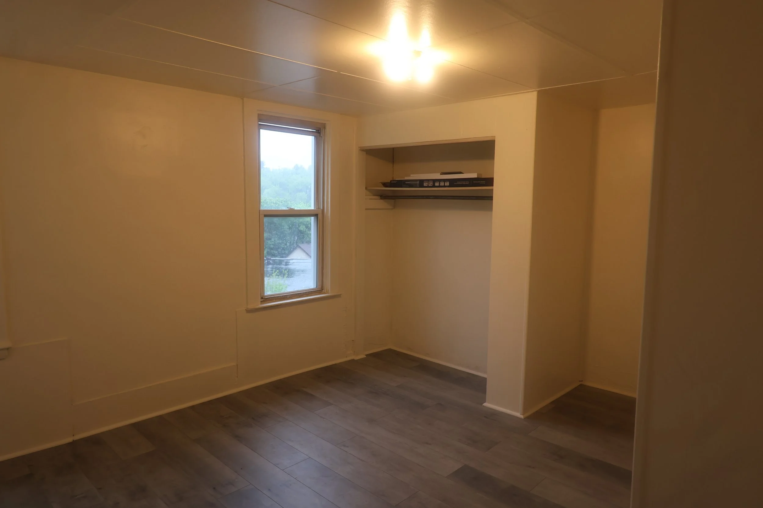 Empty room with beige walls, a window showing trees outside, a built-in wall shelf with some items, and wood flooring.