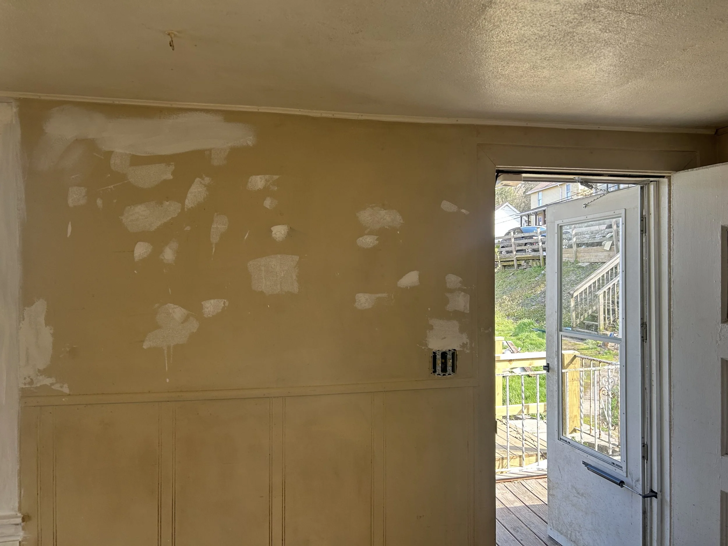 Interior of a room under renovation with a partially painted wall, an open door leading to a wooden deck, and a hillside with houses in the background.