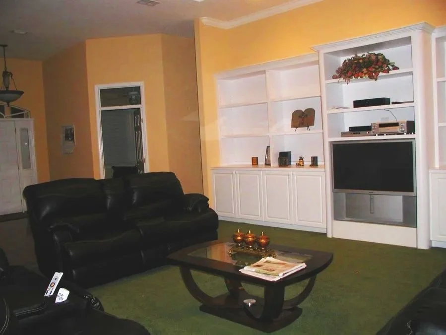 Living room with yellow walls, black leather sofas, a wooden coffee table with magazines and decorative items, a white built-in entertainment center with shelves, a television, and decorative plants.