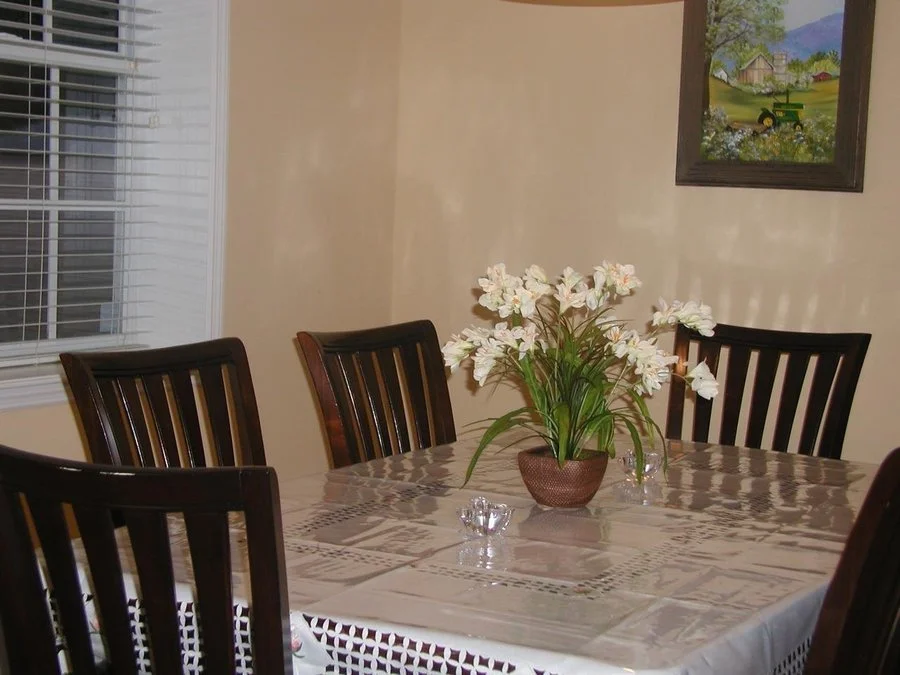Dining room table with a floral centerpiece and chairs, window with blinds, and framed landscape painting on the wall.