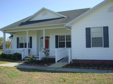 Front view of a white house with a porch, black shutters, and a small garden bed in front.