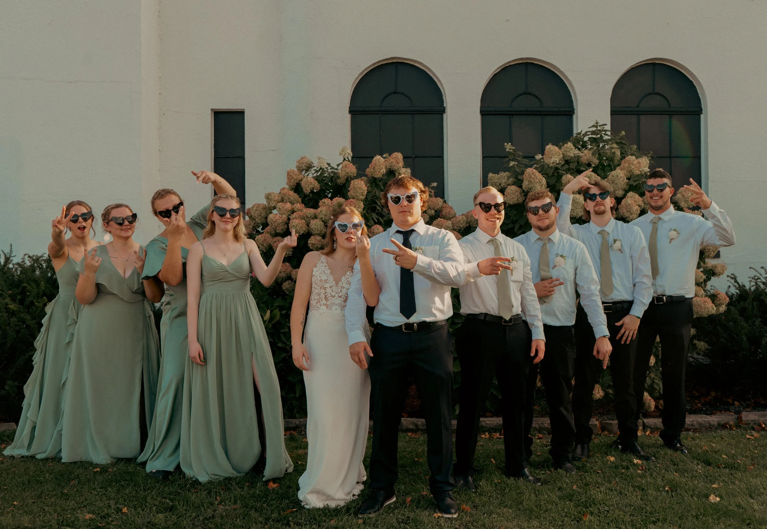 Group of wedding guests in sunglasses, posing outdoors in front of flowering bushes, some making peace signs and playful gestures.