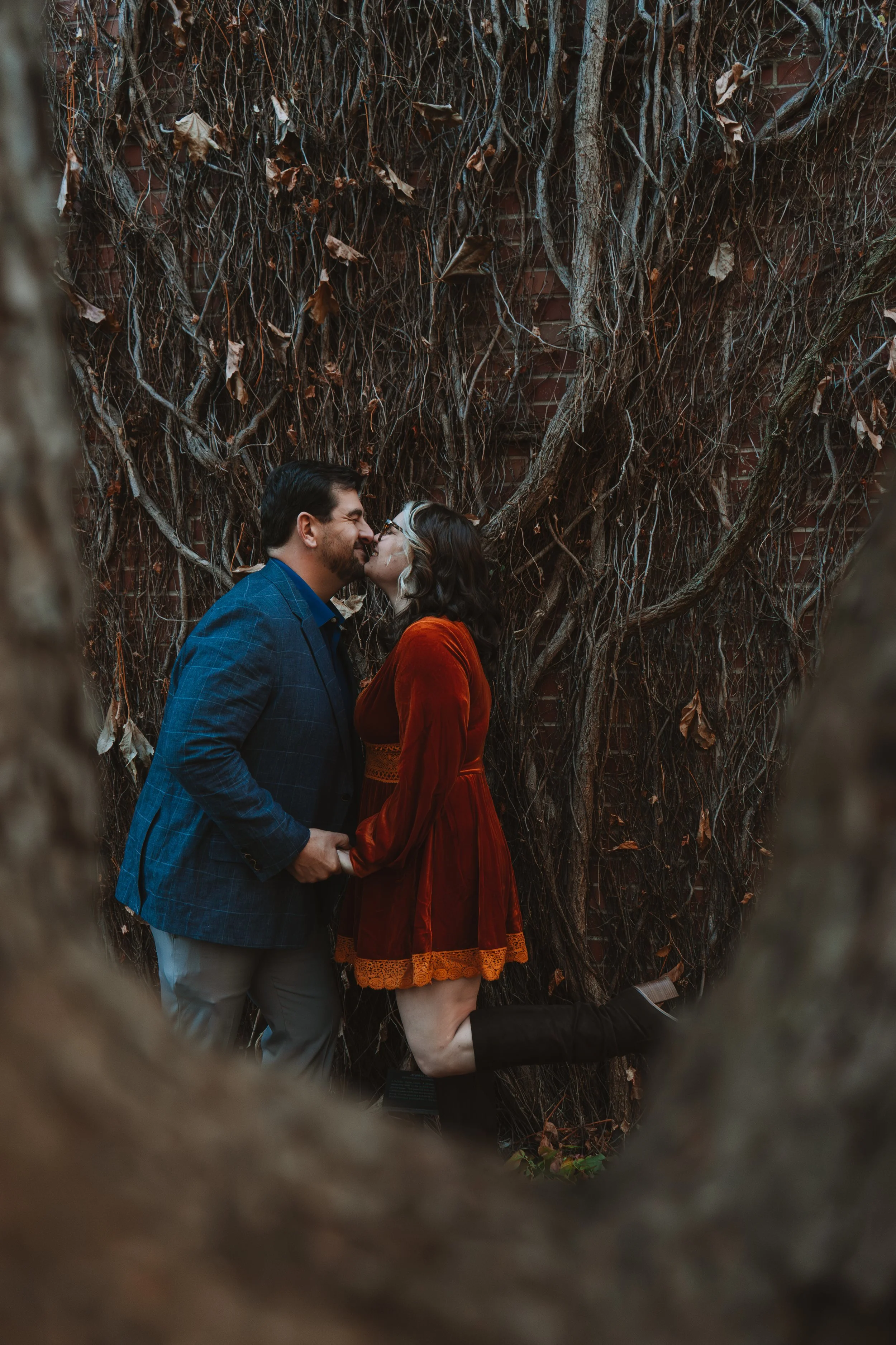 A couple kissing in front of a wall covered with tangled vines, with the woman's leg bent at the knee and wearing black knee-high boots.