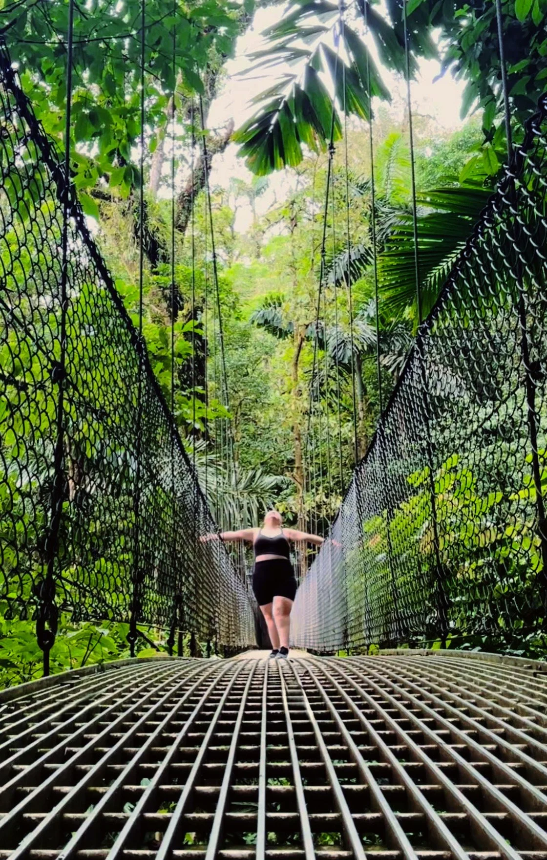 A woman in a black sports bra and shorts walking on a metal suspension bridge in a dense green jungle.
