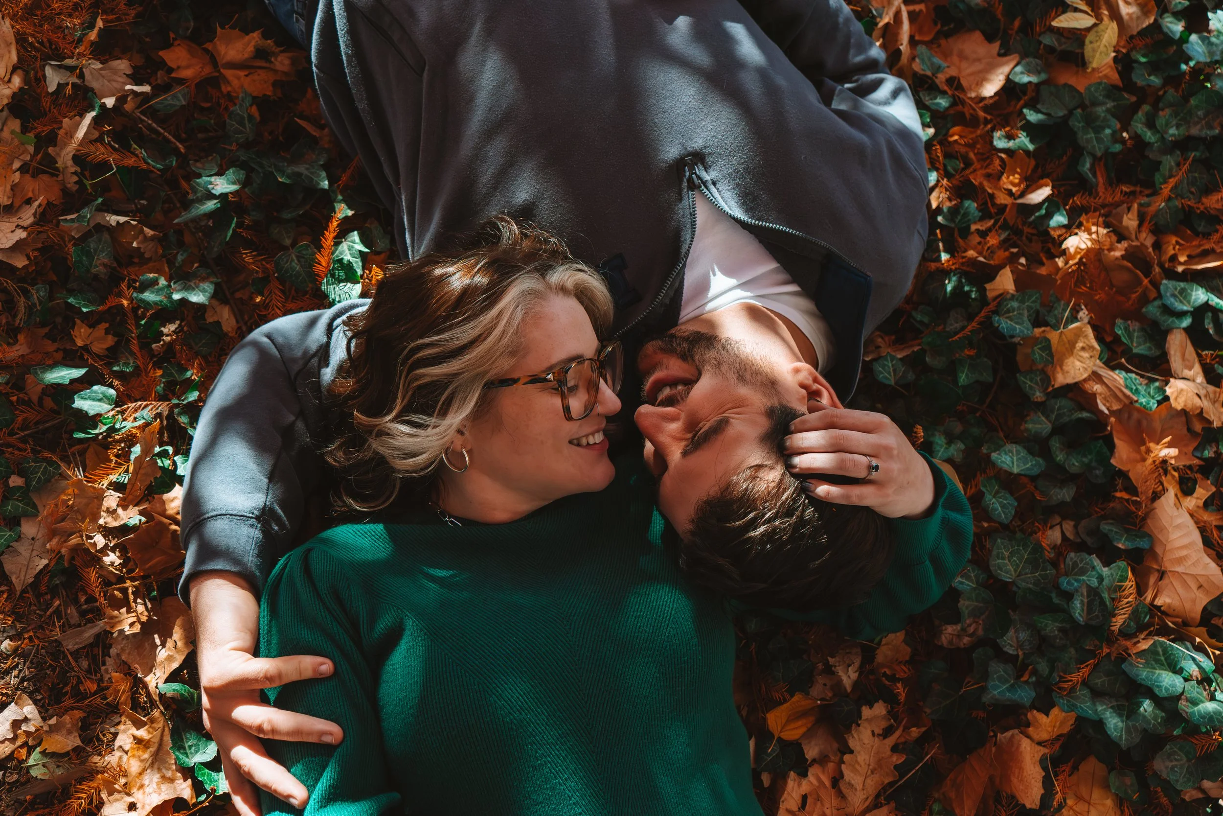 A couple lying on the ground covered in autumn leaves, smiling and looking at each other, as the woman gently touches the man's face.