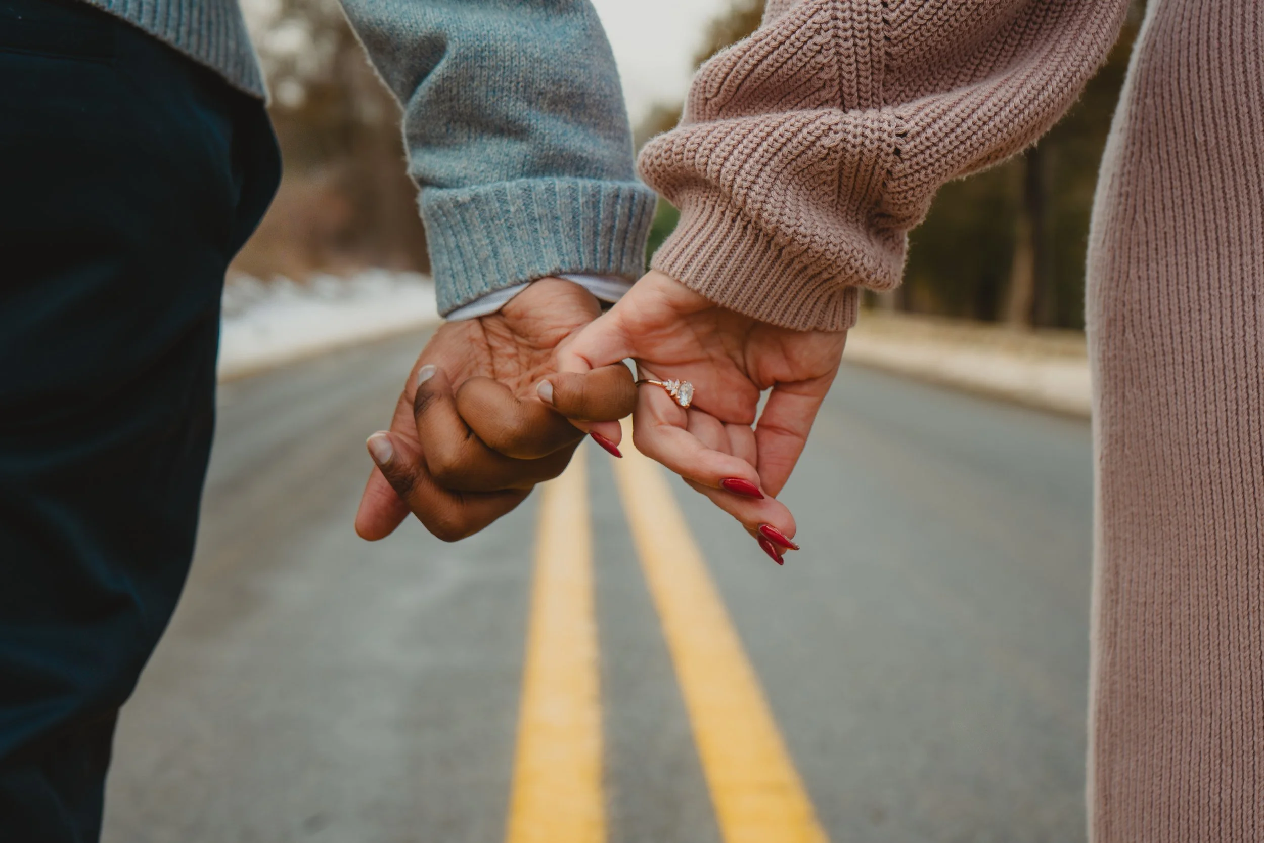 A close-up of a couple holding hands with their fingers intertwined, walking on a double yellow line road with a wintery background.