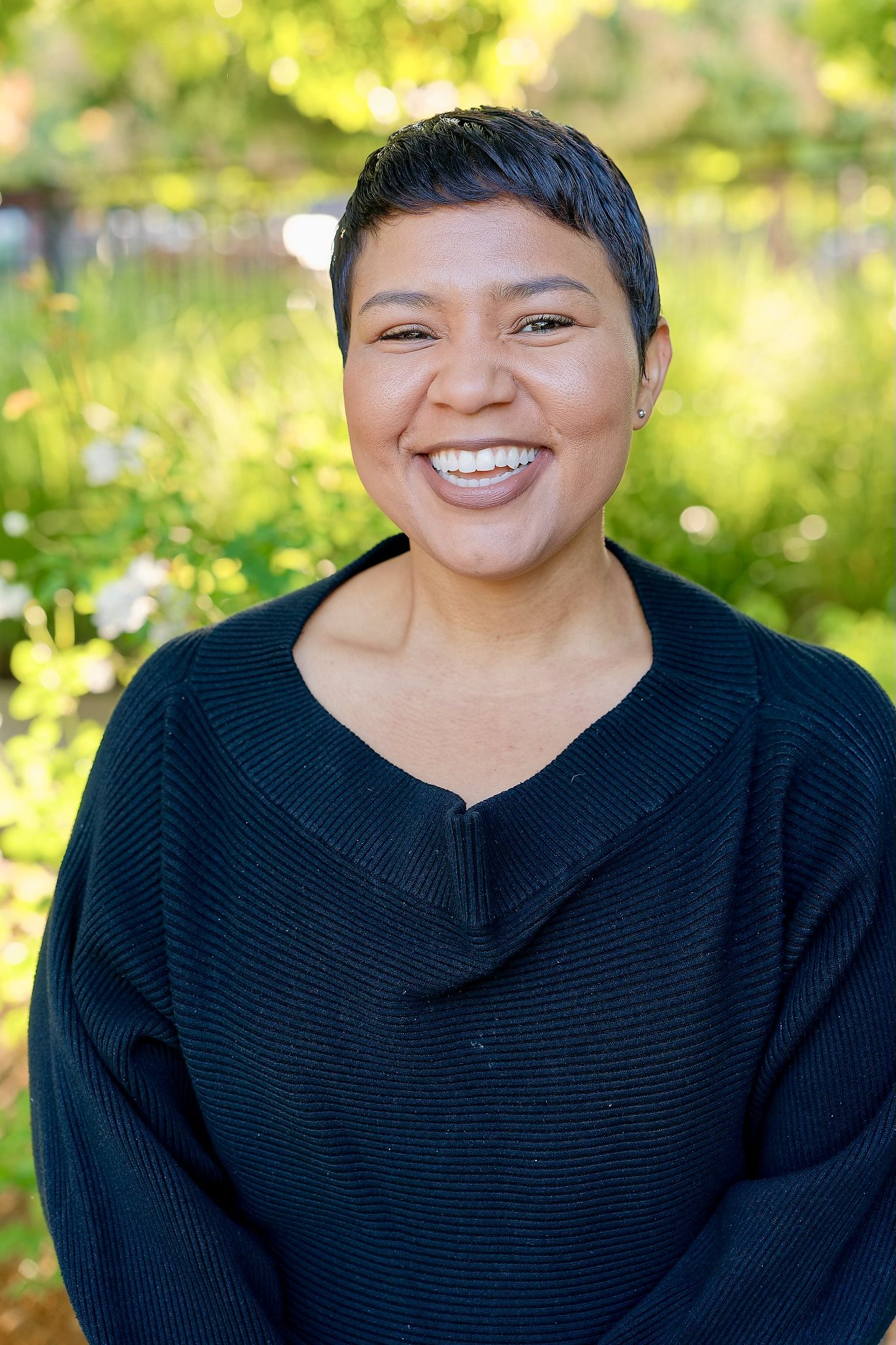 A woman with short dark hair smiling and winking outdoors in front of green foliage, wearing a black sweater.