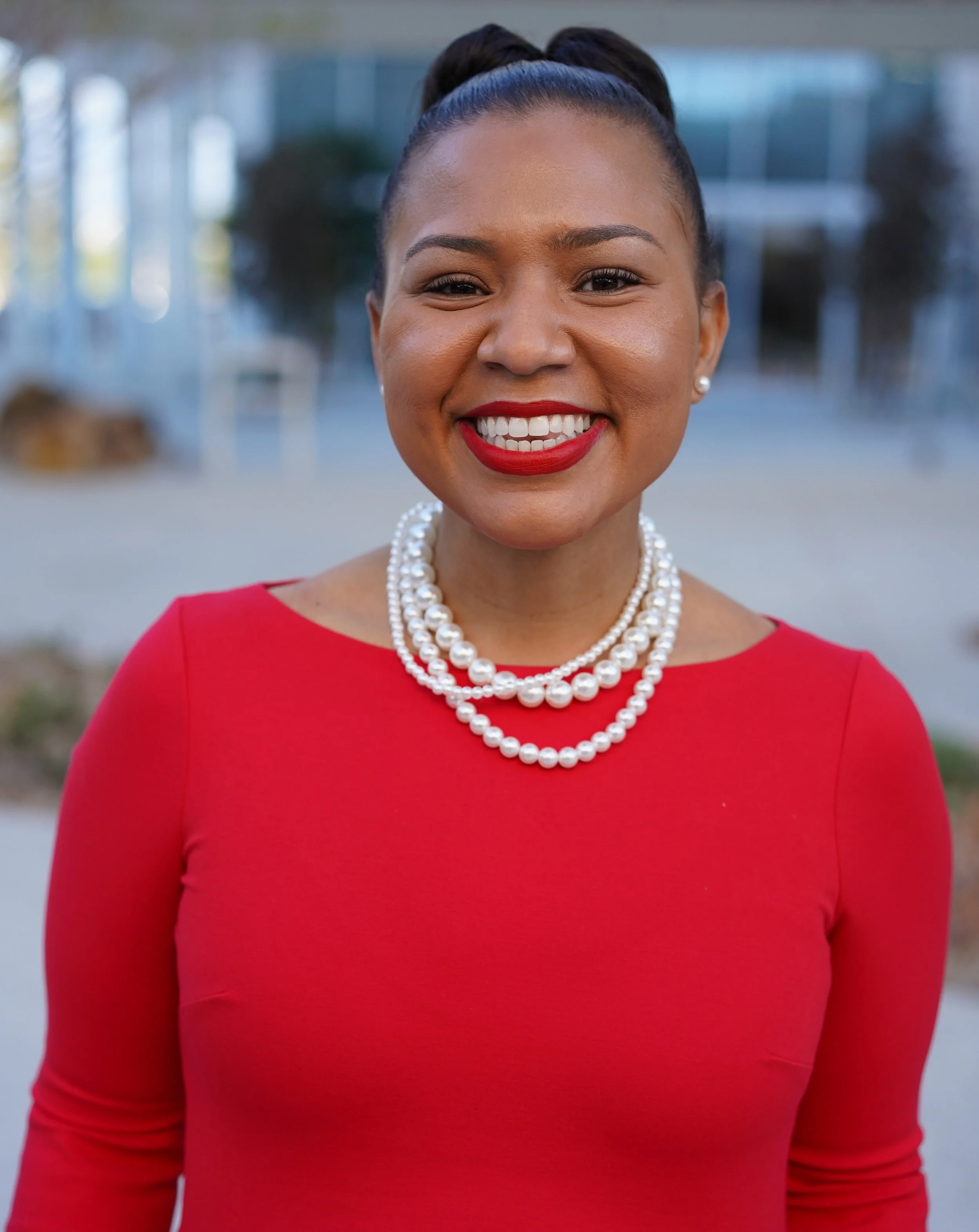Front view of a smiling woman wearing a red dress, pearl necklace, pearl earrings, and a high bun hairstyle, outdoors with blurred background of buildings and trees.
