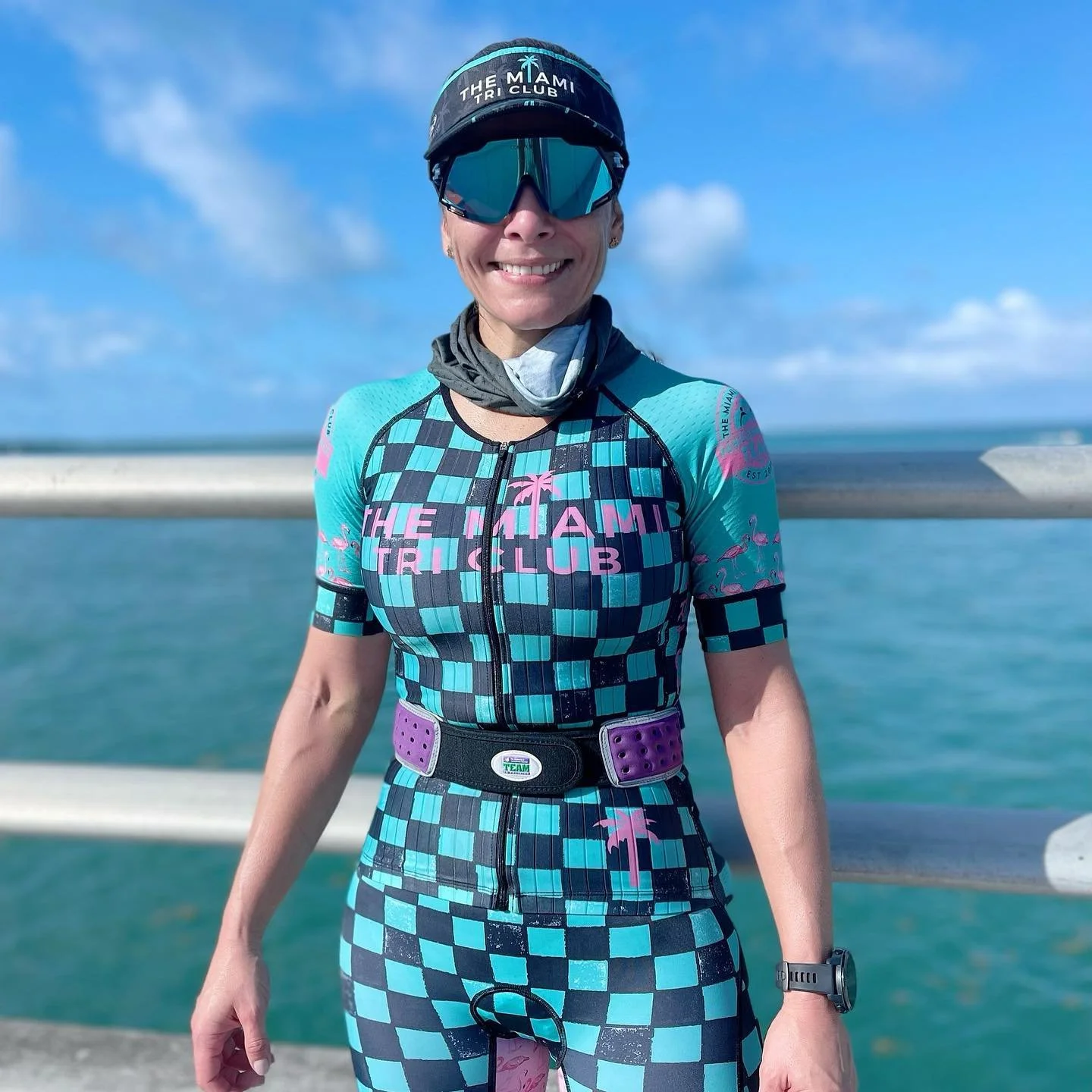 A woman dressed in a colorful cycling outfit with the words 'The Miami Tri Club' on her jersey, standing on a pier near the ocean, wearing sunglasses and a cap, smiling at the camera.