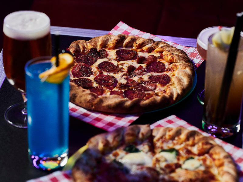 Two pizzas on red and white checkered napkins with drinks, including a beer, a blue cocktail, and a dark soda with a straw, on a black table.