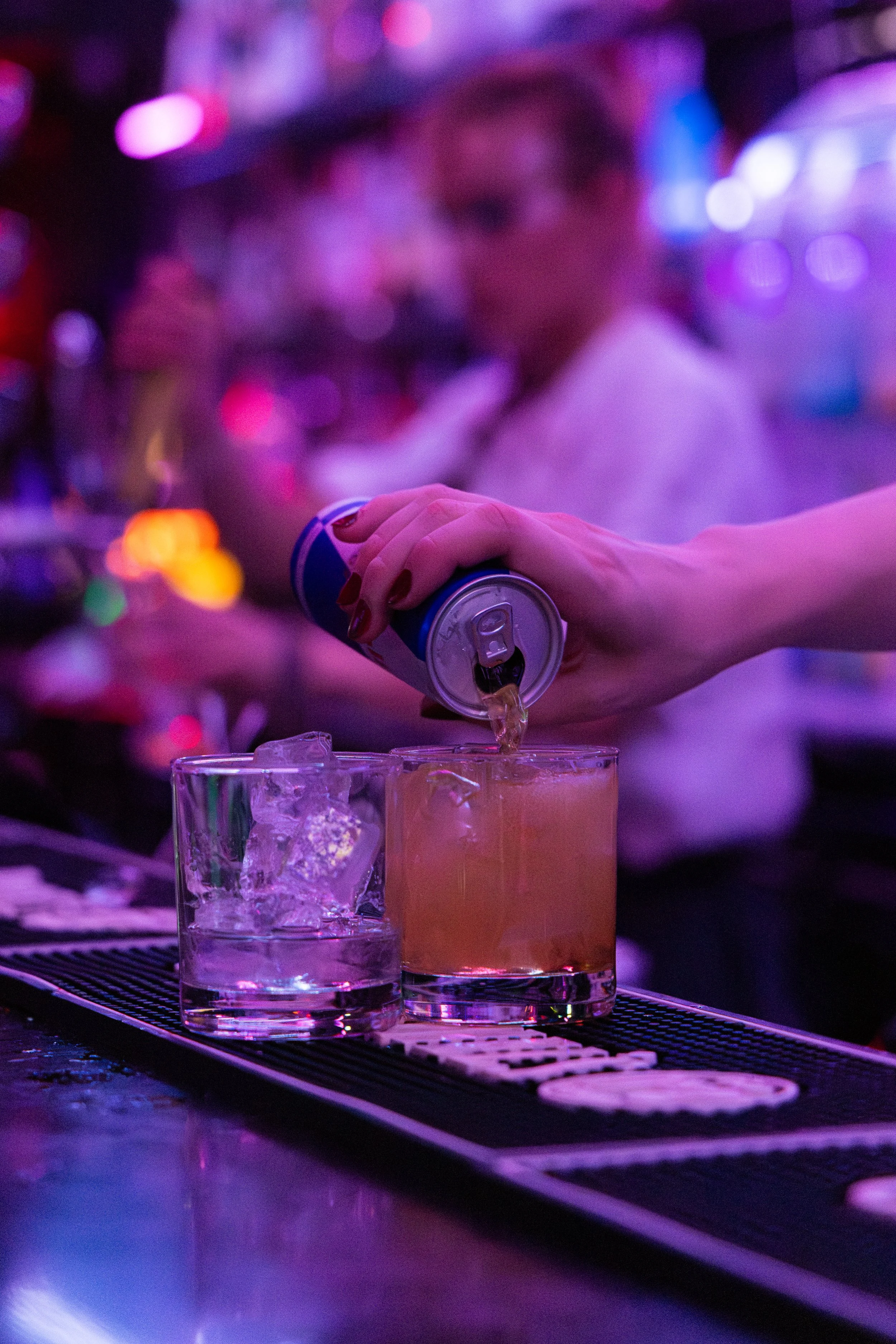 A person pouring a drink from a can into a glass at a bar or nightclub with colorful, blurred neon lights in the background.