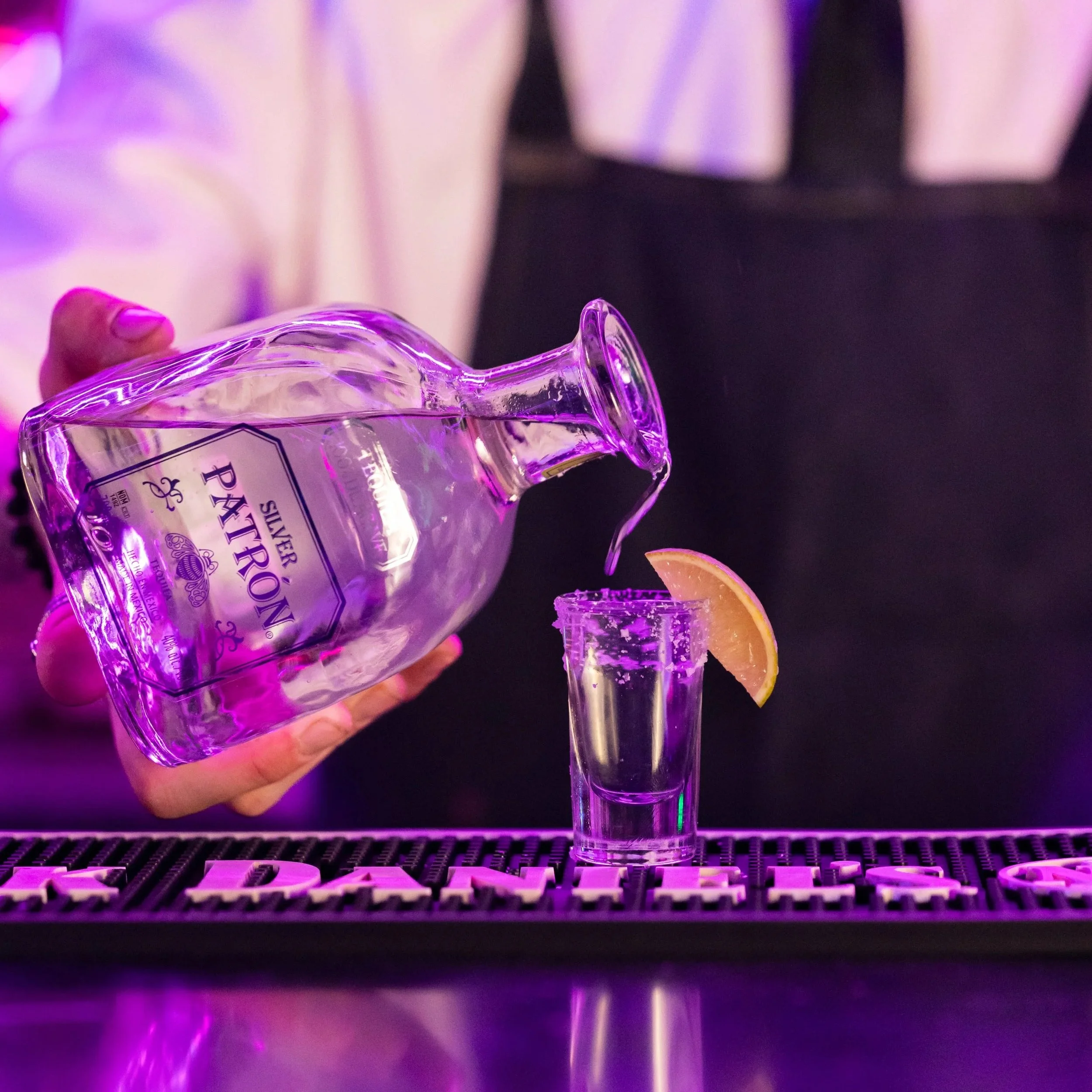 A person pouring pink-colored liquid from a bottle into a shot glass, with a lemon wedge on the counter and purple lighting giving the scene a vibrant, nightlife atmosphere.