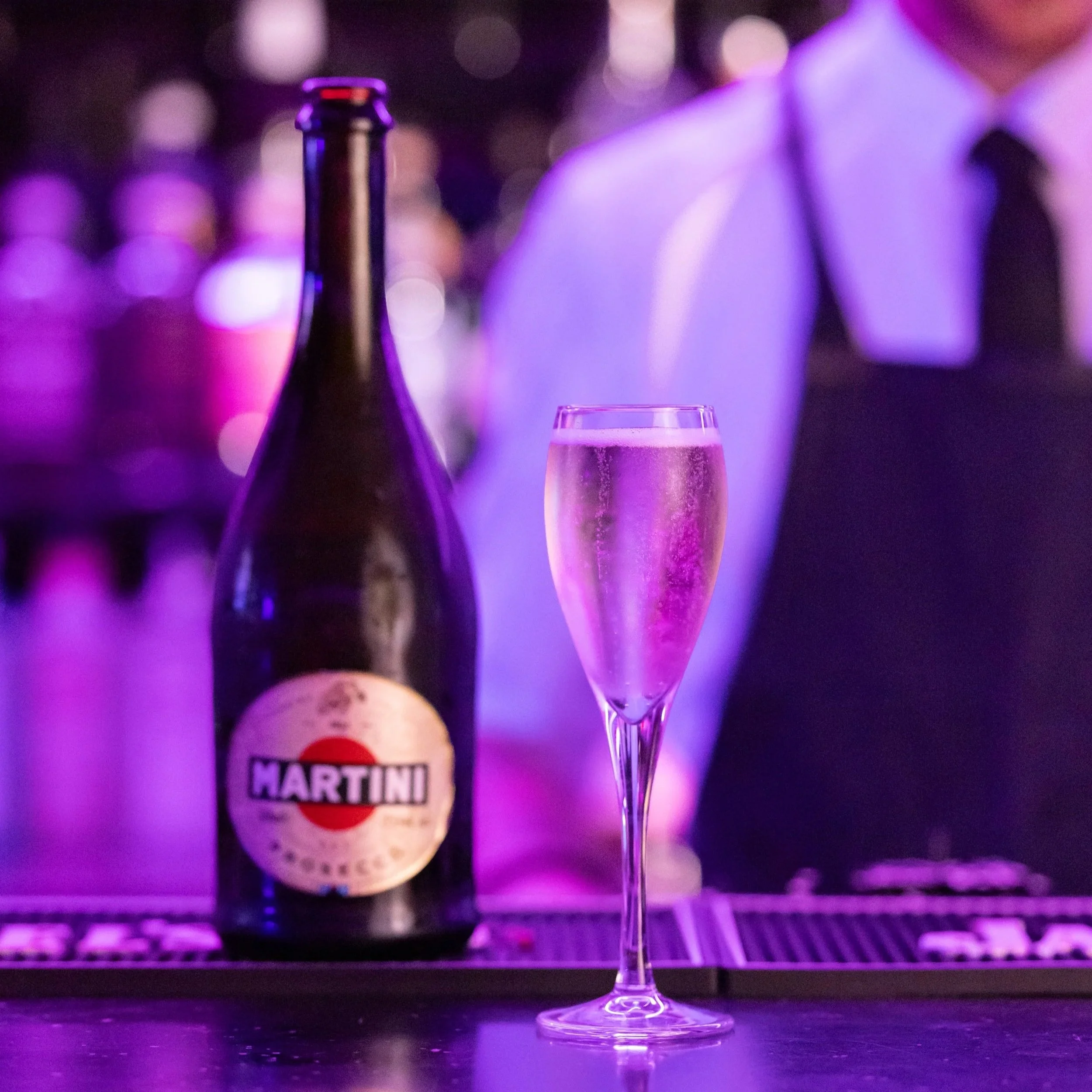 A glass of champagne, a champagne bottle, and a bottle labeled 'Martini' are arranged on a dark surface with colorful bokeh lights in the background.