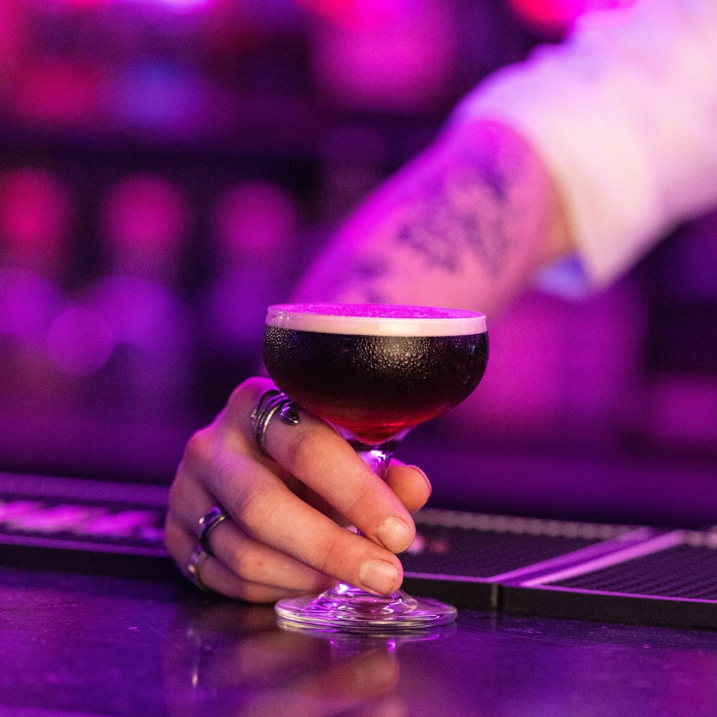 Close-up of a hand holding a glass with purple-colored drink, with blurred purple and pink lights in the background.