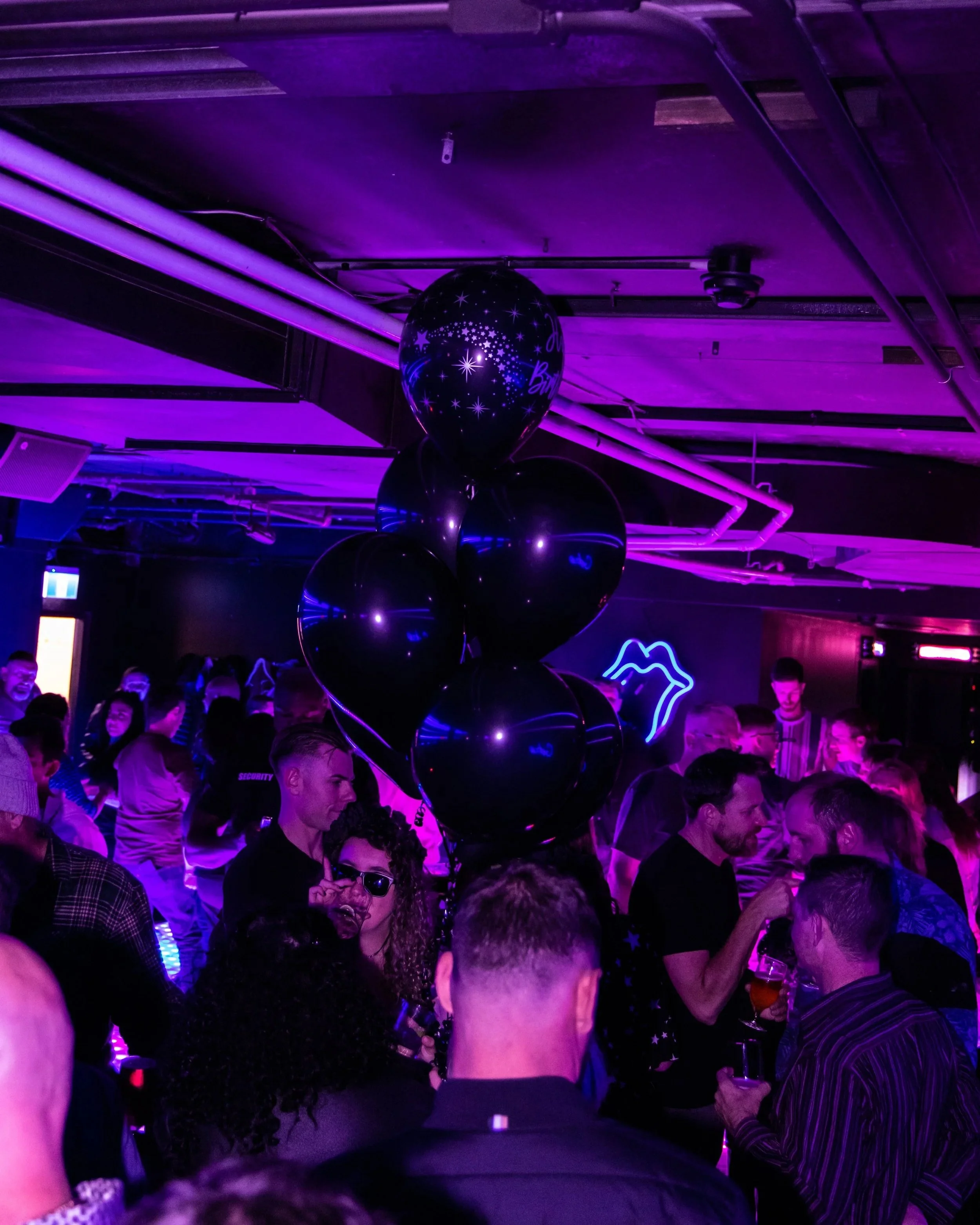 A nightclub or party scene with a crowd of people under purple and blue lighting. Black balloons with star designs float above the crowd. Neon signs are visible on the wall, and some individuals are holding drinks.