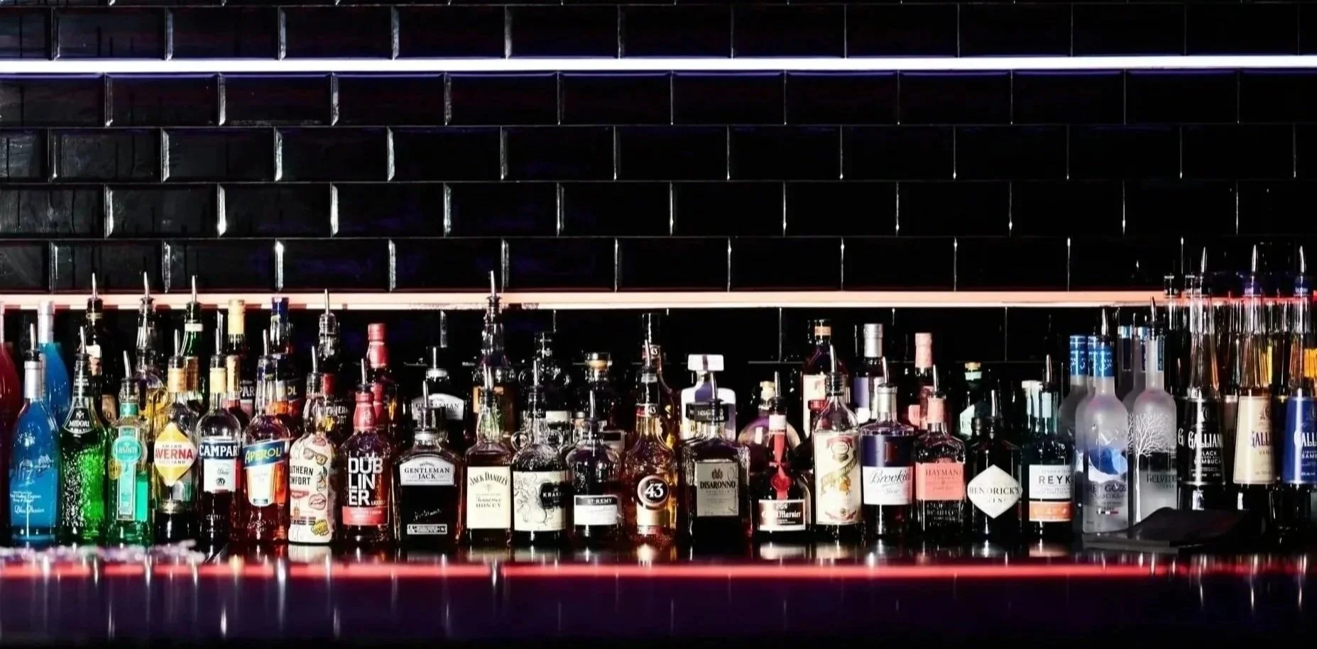 Shelf filled with various bottles of alcohol behind a bar, against a black tiled wall with a white neon light stripe.