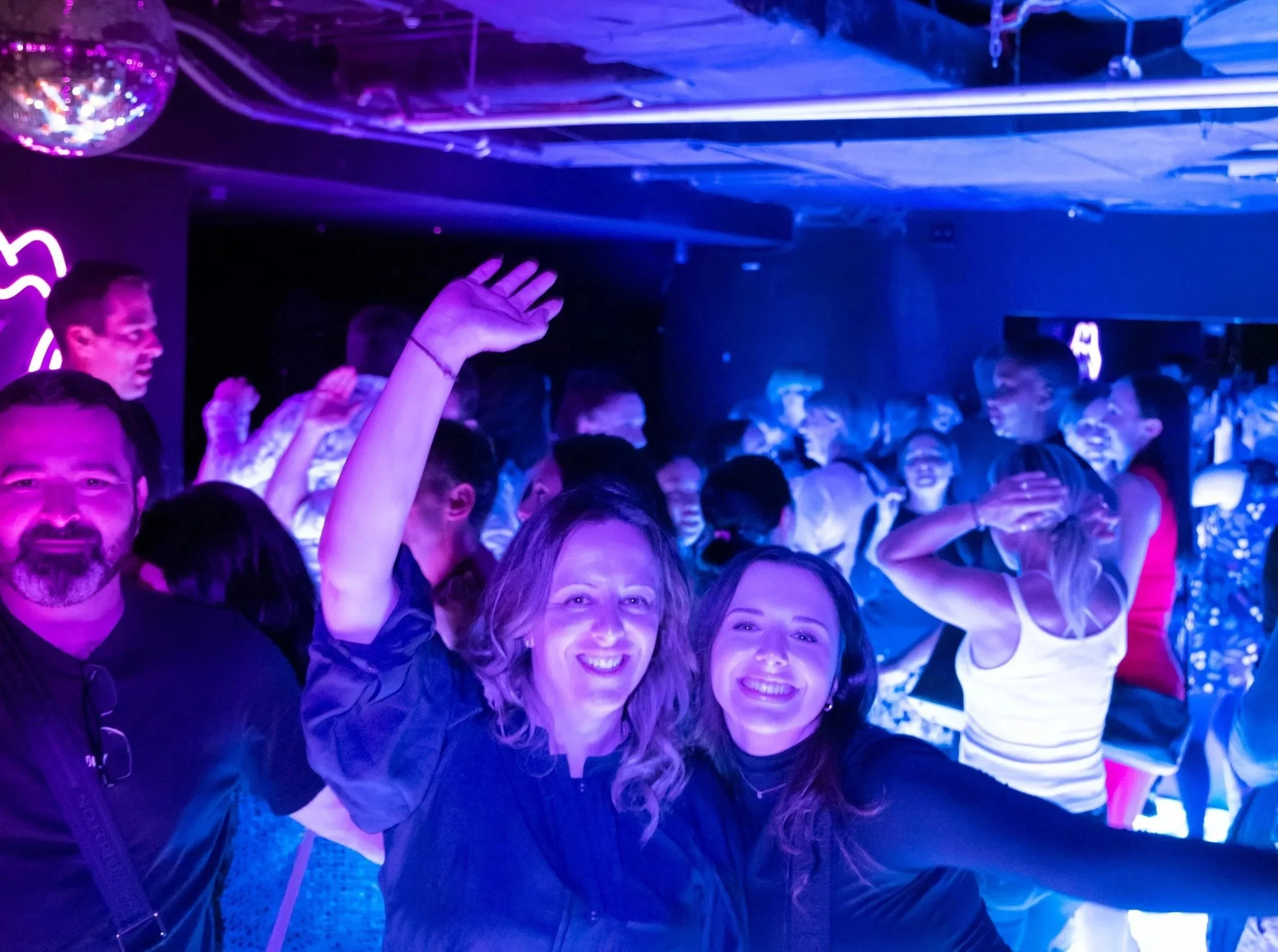 A crowded party with people dancing under purple and blue lighting, with some smiling and raising their hands.