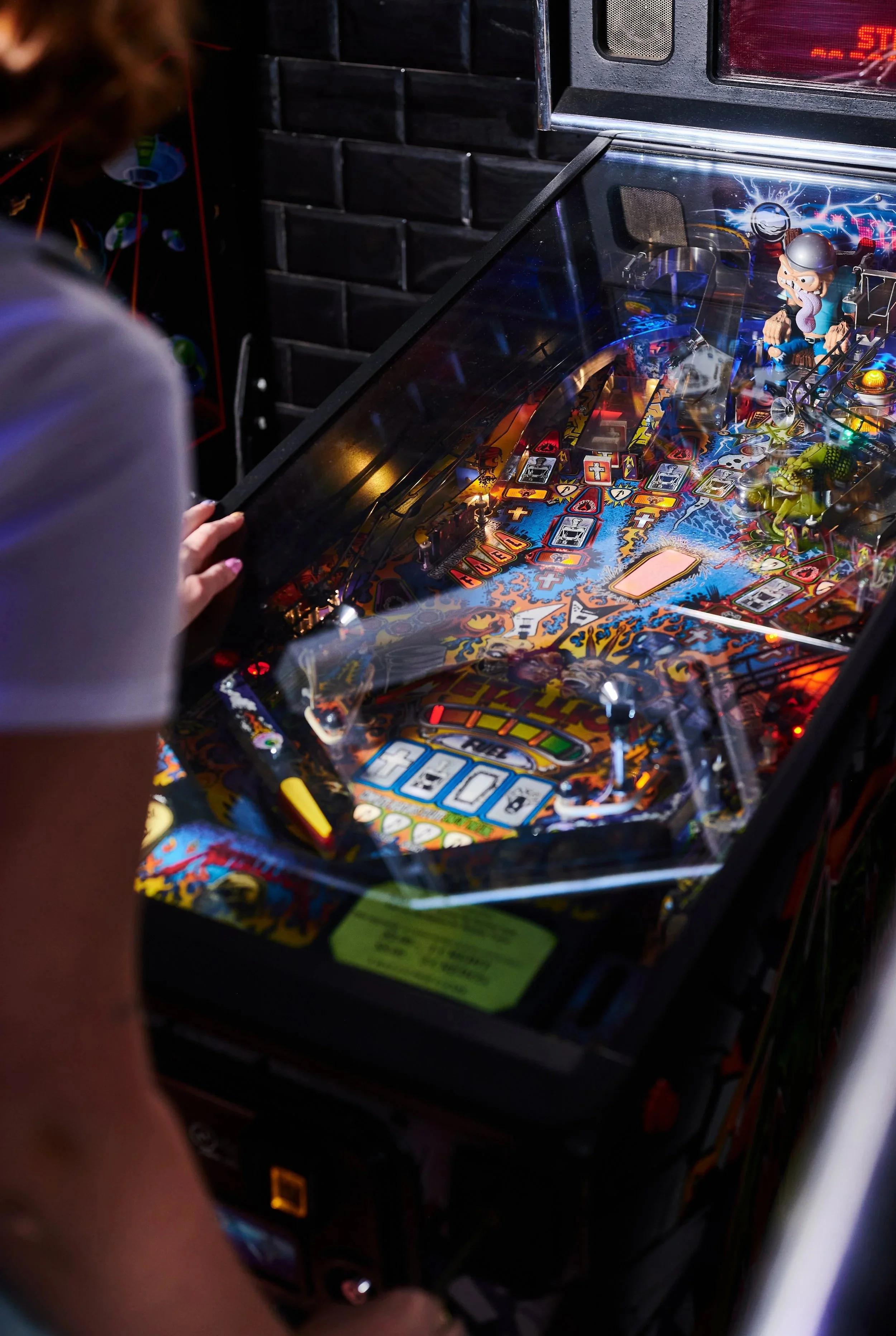 Close-up of a pinball machine with colorful artwork, illuminated lights, and a cartoon character toy on top. A person with a hand near the machine is visible on the left side, with a brick wall in the background.