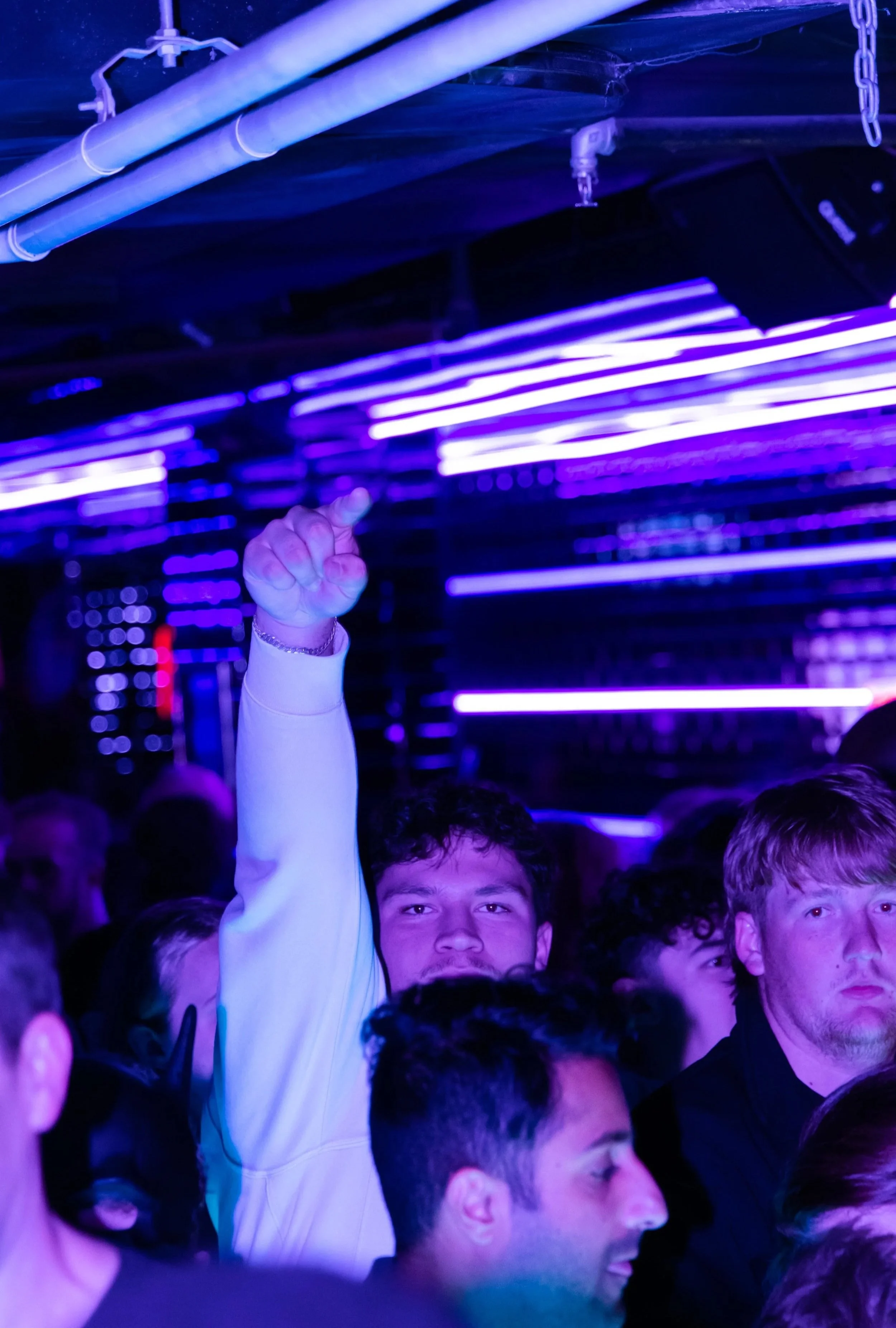 A group of young people in a nightclub or concert setting with purple neon lights overhead. One person in the crowd is raising their hand with a finger pointing upwards.
