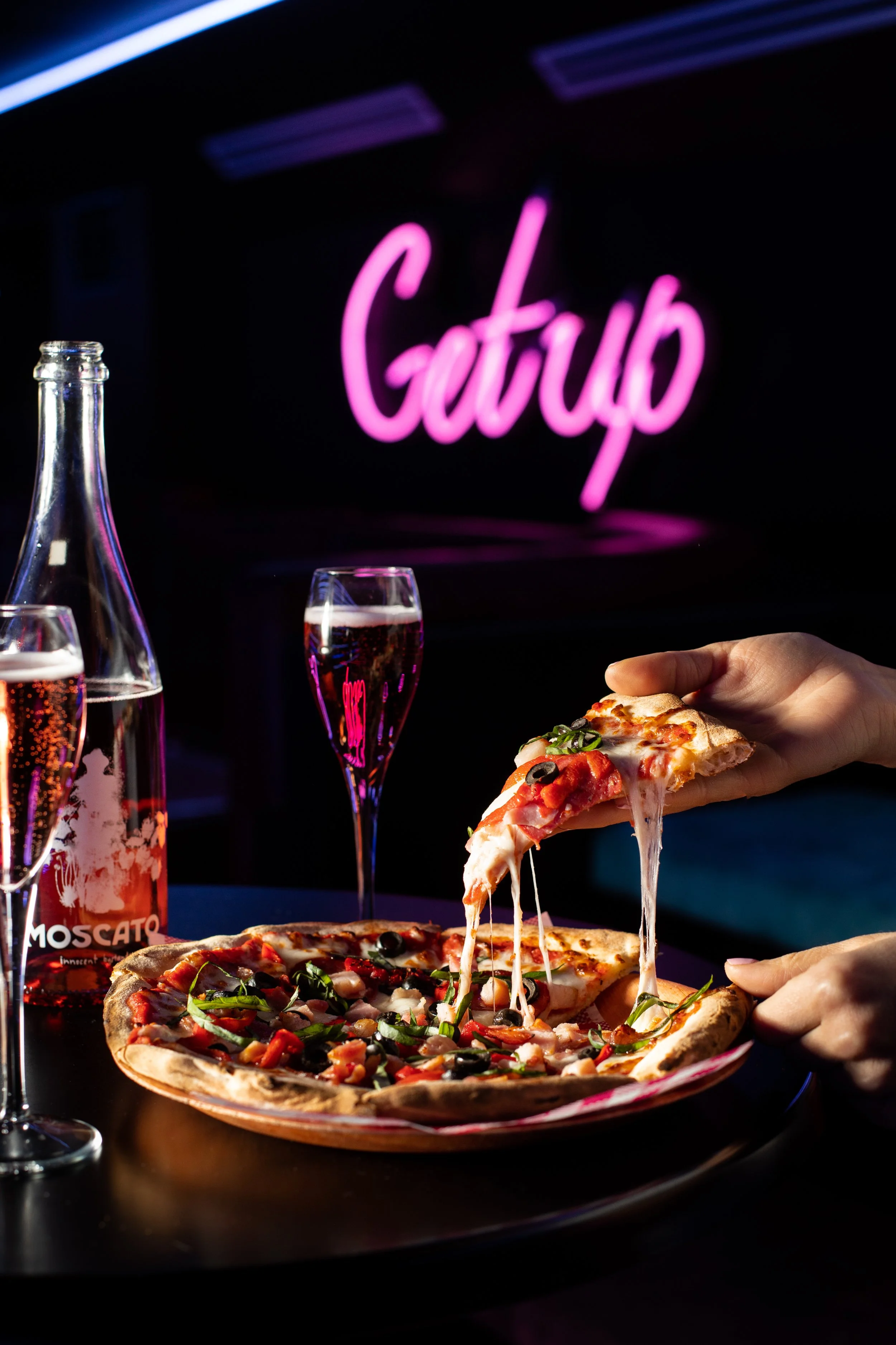 Close-up of a person serving a slice of pizza with toppings of olives, peppers, and greens onto a plate. A bottle of Moscato wine, two glasses of sparkling wine, and a neon pink sign that says 'Gatub' are visible in the background.