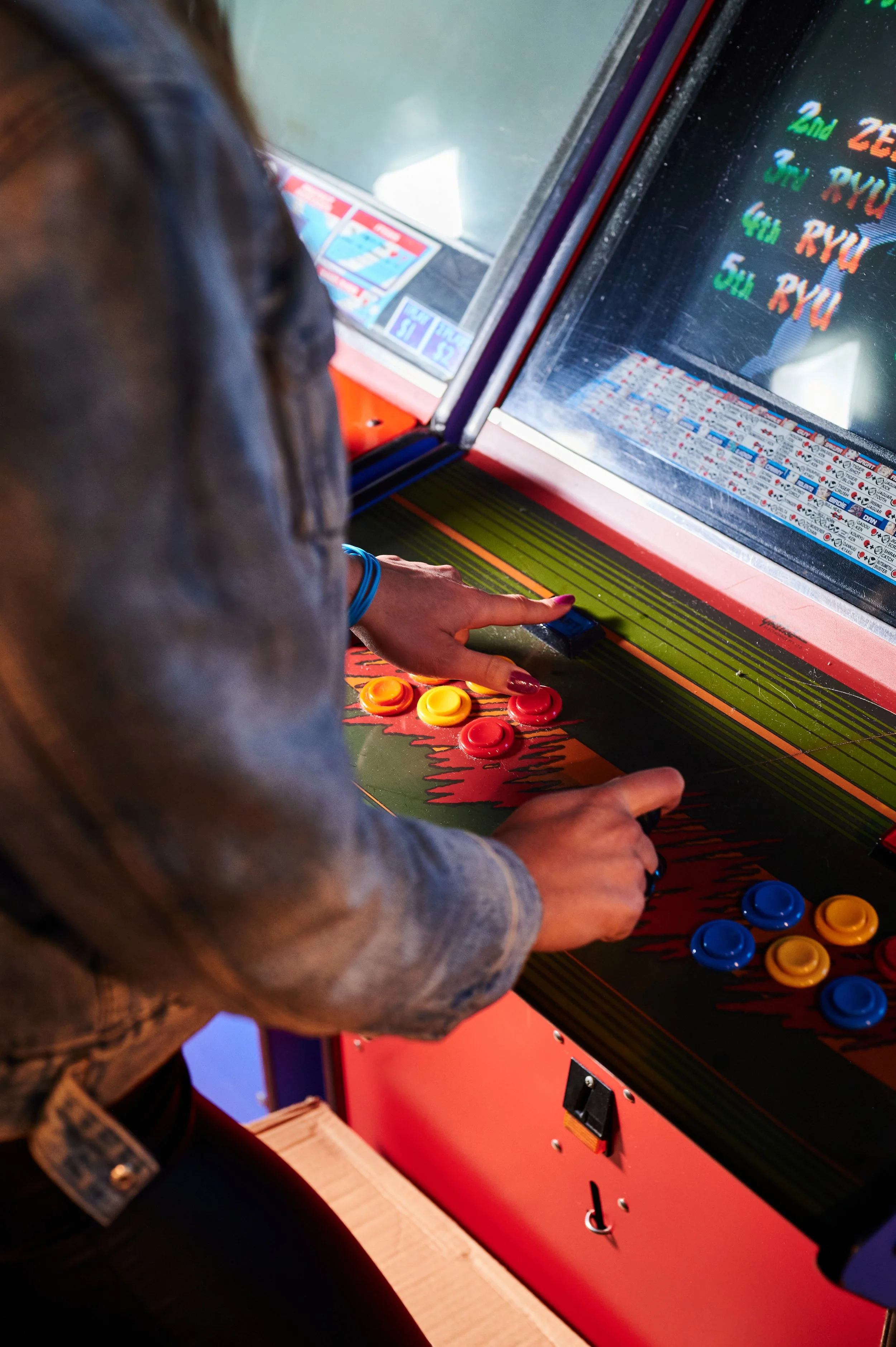 Close-up of a person playing a vintage arcade game, pushing a yellow button with their right hand while their left hand presses a red button, with colorful poker chip tokens scattered on the game console.