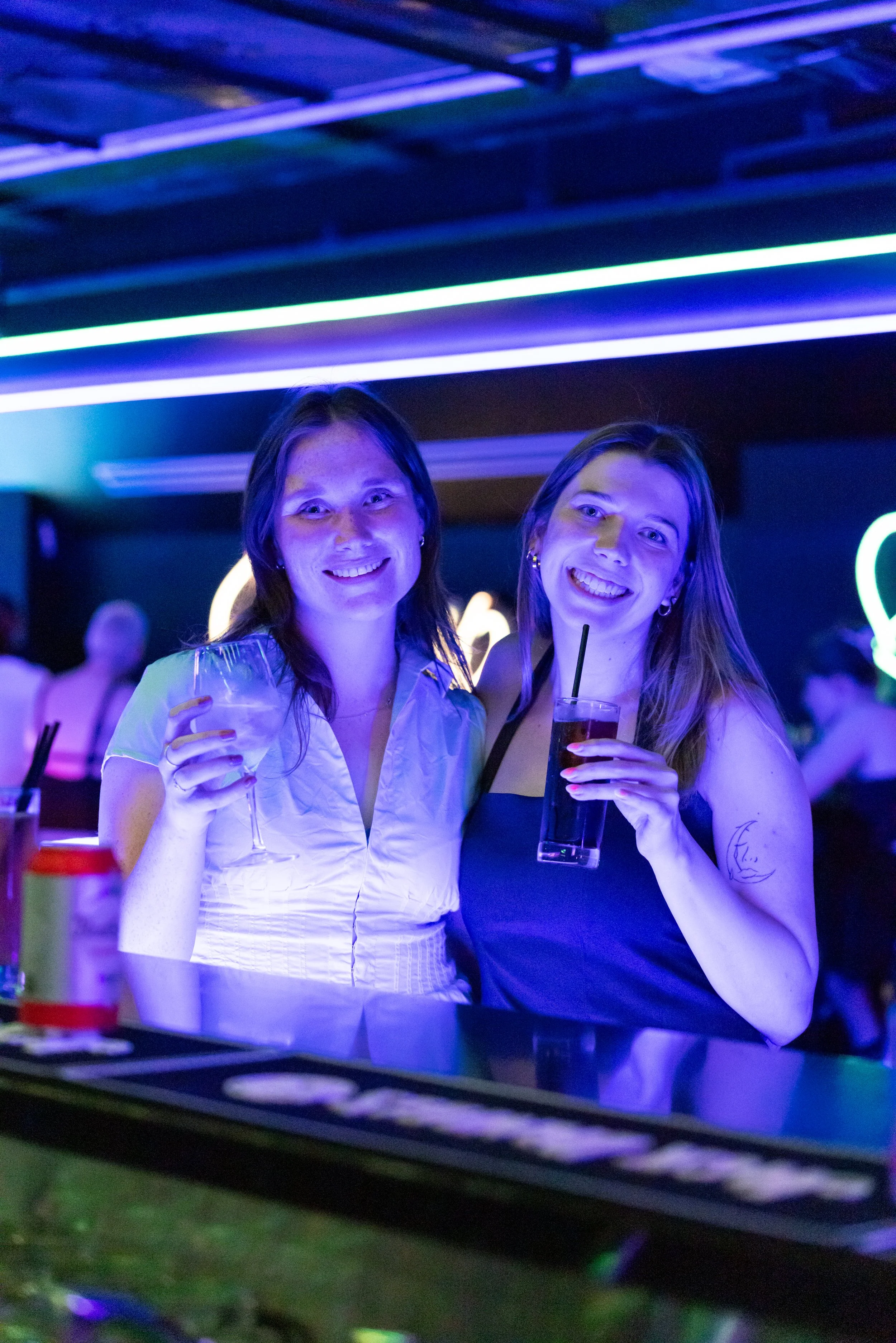 Two women smiling and holding drinks at a bar or club with neon lighting.