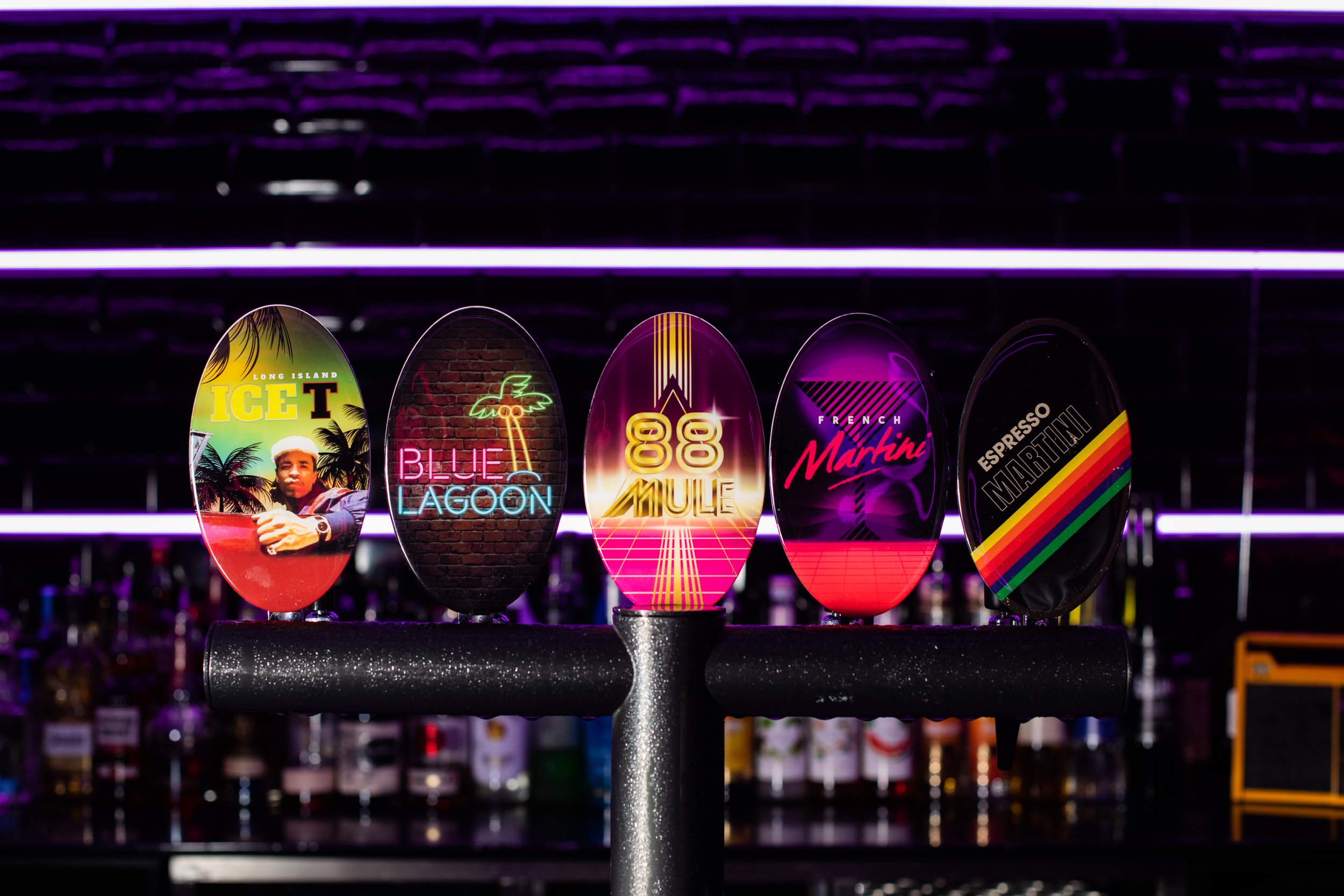 A display of colorful neon drink signs on a stand, with each sign showing different designs and text, including 'Long Island Ice T,' 'Blue Lagoon,' '88 Mule,' 'French Martini,' and 'Espresso Martini,' illuminated against a dark background.