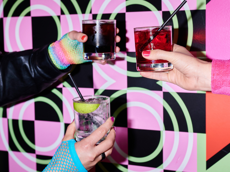 Three hands holding colorful cocktails over a patterned background