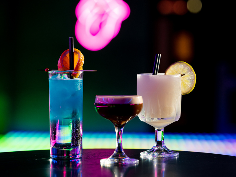 Three colorful cocktails on a bar counter with vibrant neon lights in the background.
