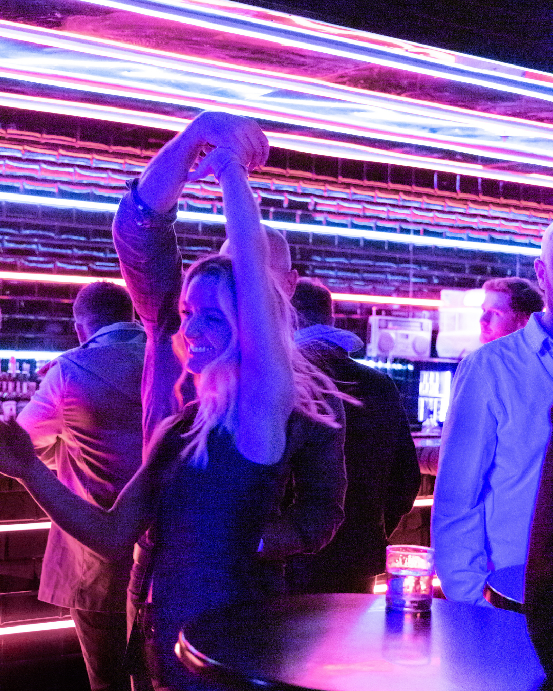 A woman dancing and smiling in a bar with neon purple and pink lights, surrounded by other people.