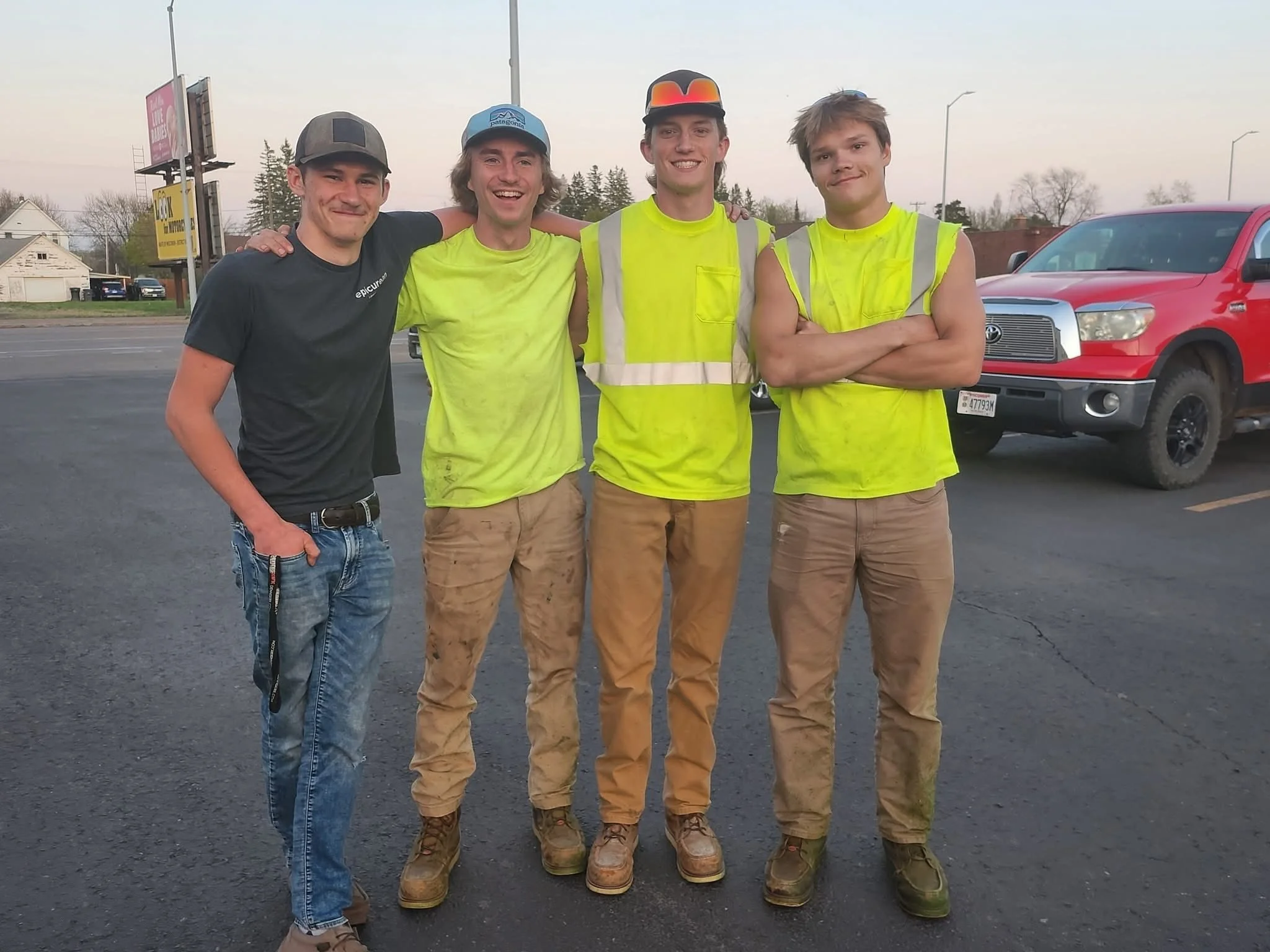 Four young men standing together in a parking lot, smiling, with two of them wearing reflective yellow safety vests. There is a red pickup truck in the background and a billboard on the side of the road.