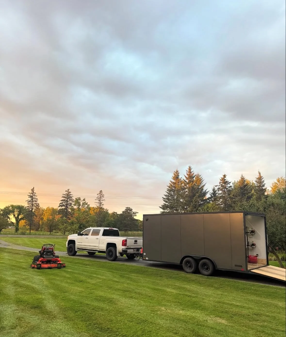 A white pickup truck with a black trailer attached parked on a grassy field, a riding lawn mower nearby, and trees in the background under a cloudy sky.