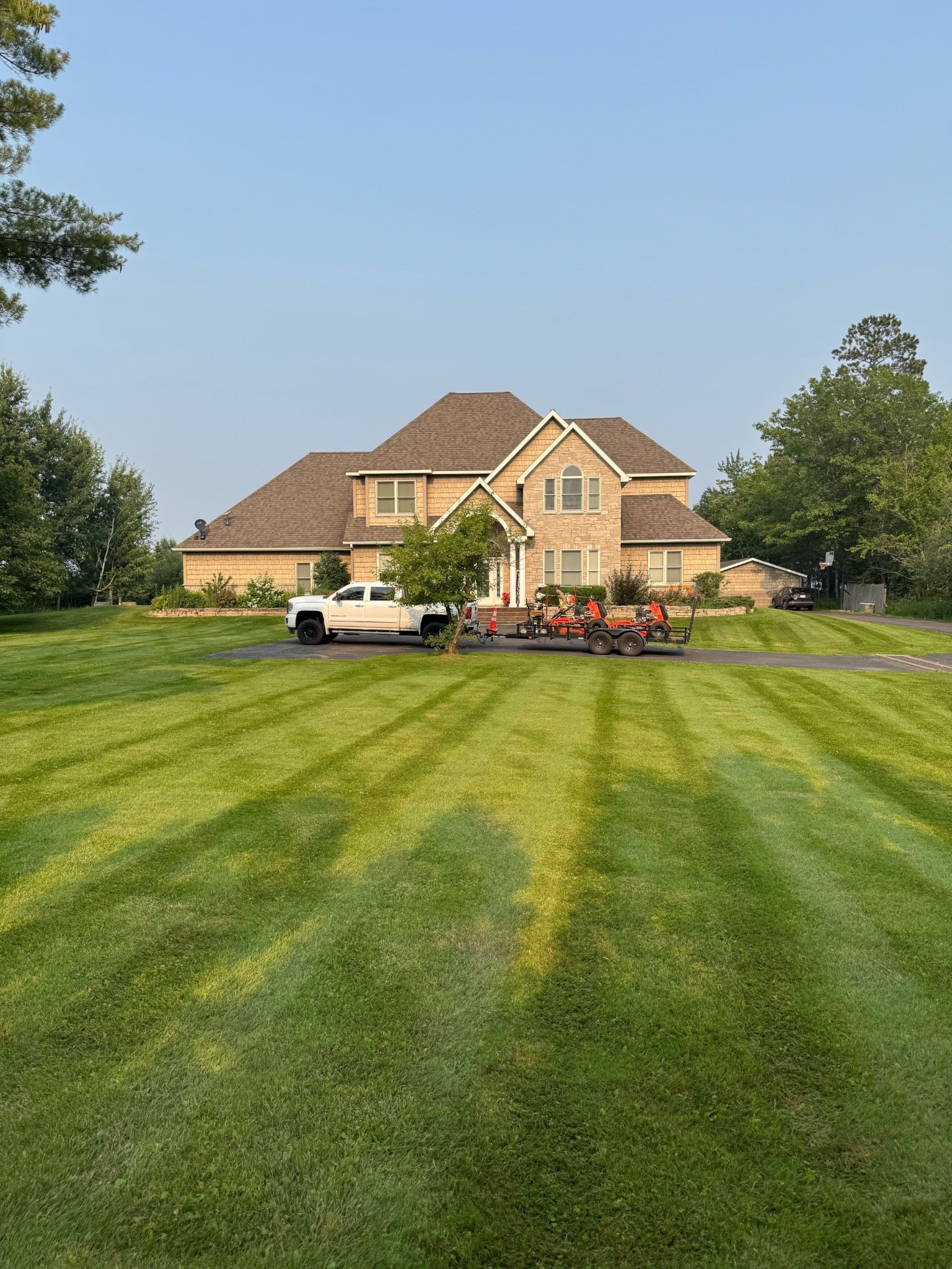 Large two-story brick house with a manicured lawn in the foreground, parked white truck and trailer with riding lawnmowers, trees on either side, and a clear blue sky.