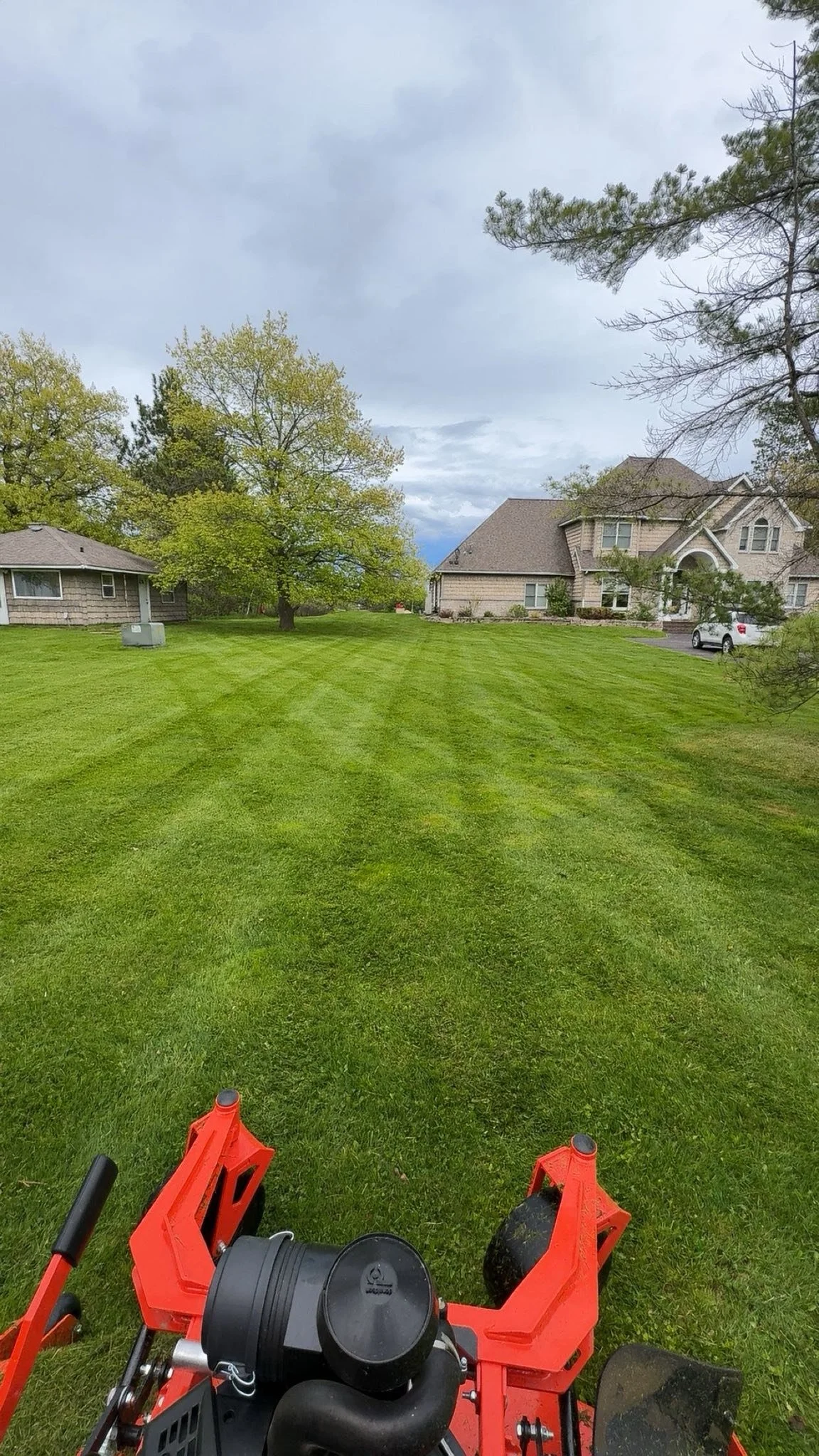 View of a well-maintained green lawn with striped mowing lines, houses in the background, trees, and cloudy sky overhead, taken from behind a lawnmower.