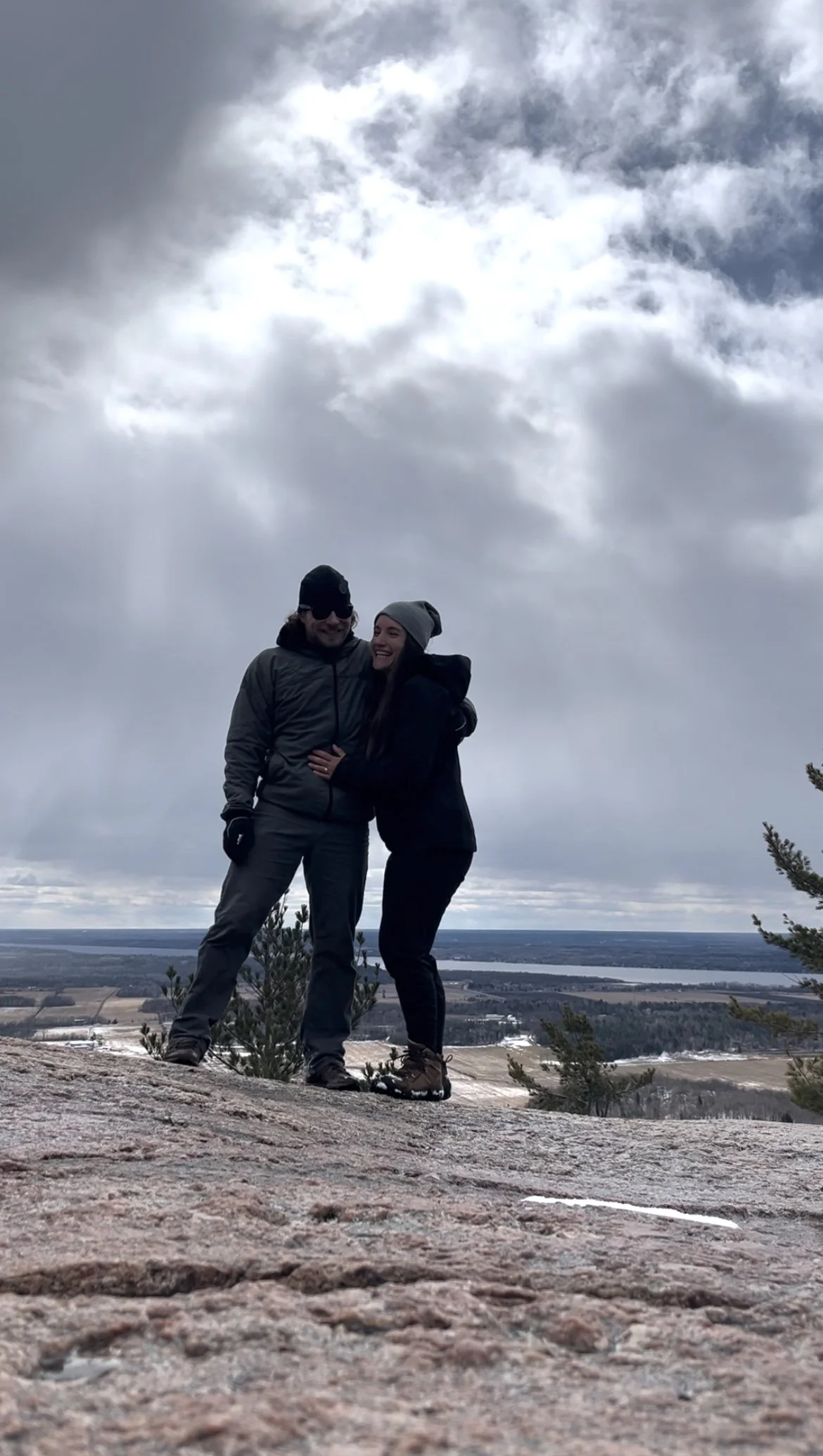A couple standing on a rocky hilltop with a cloudy sky and a scenic landscape in the background, dressed in winter clothing.