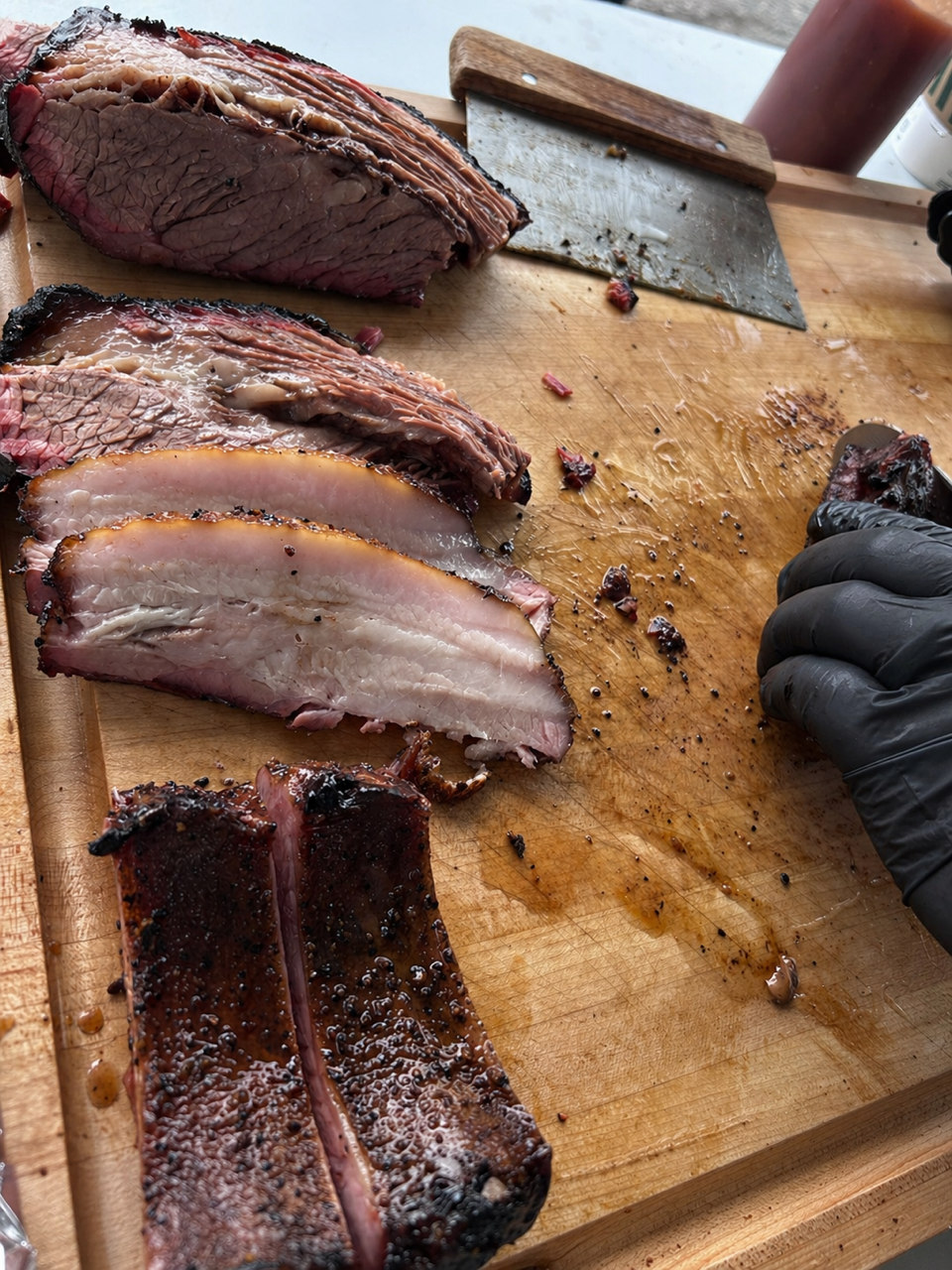 Slices of smoked brisket on a wooden cutting board, with a butcher's knife, a bottle of barbecue sauce, and a person's hand wearing a black glove.