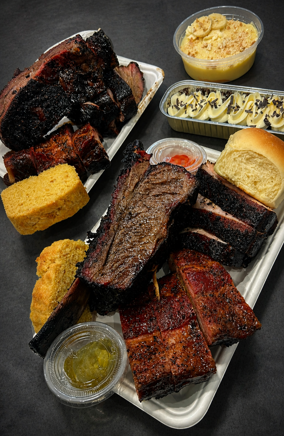 A tray of barbecued ribs, brisket, and sausage served with cornbread, bread roll, sides of mac and cheese, banana pudding, and other desserts, with dipping sauces.