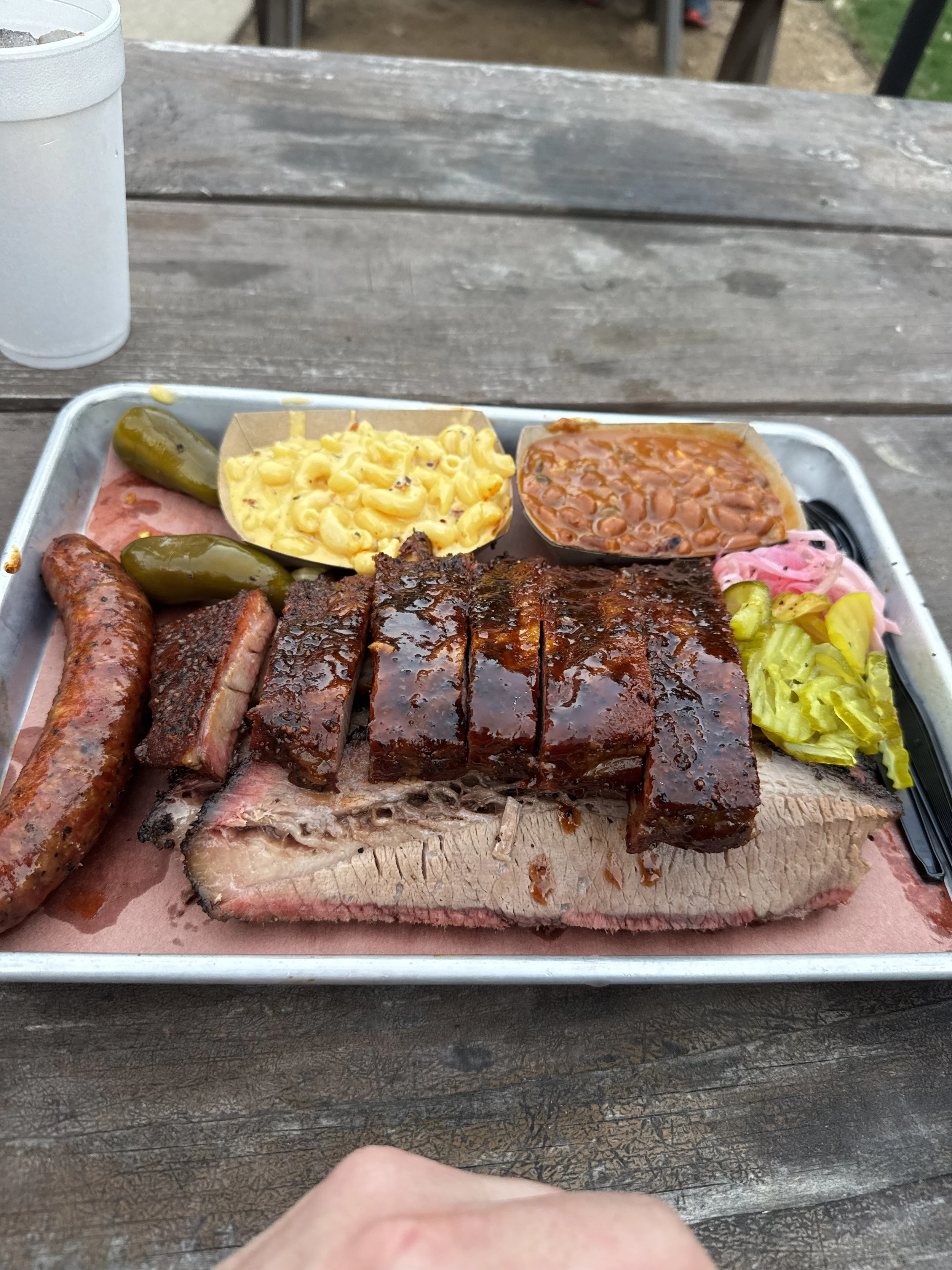 A tray of barbecue food on a wooden table containing sliced brisket, a sausage, macaroni and cheese, baked beans, pickles, and coleslaw.