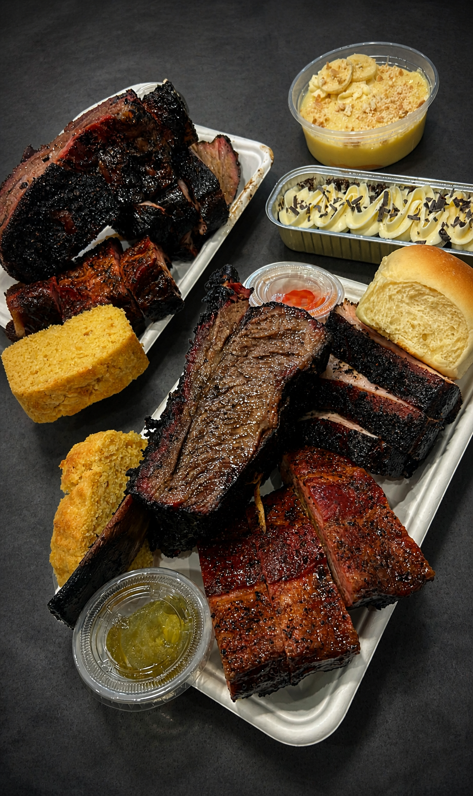 Assorted grilled and smoked meats, cornbread, and side dishes on white trays, including salad and dessert, on a black table.