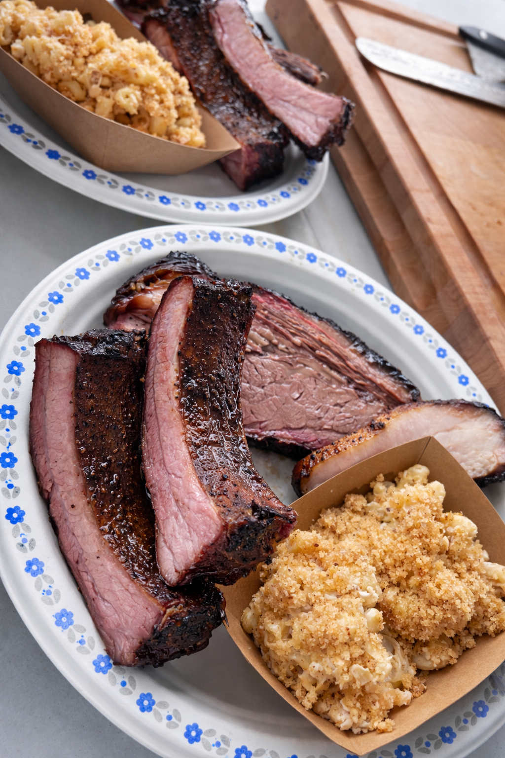 Barbecue ribs with a side of macaroni and cheese on a white plate with blue flowers. A chopping board with a knife and barbecue sauce in the background.