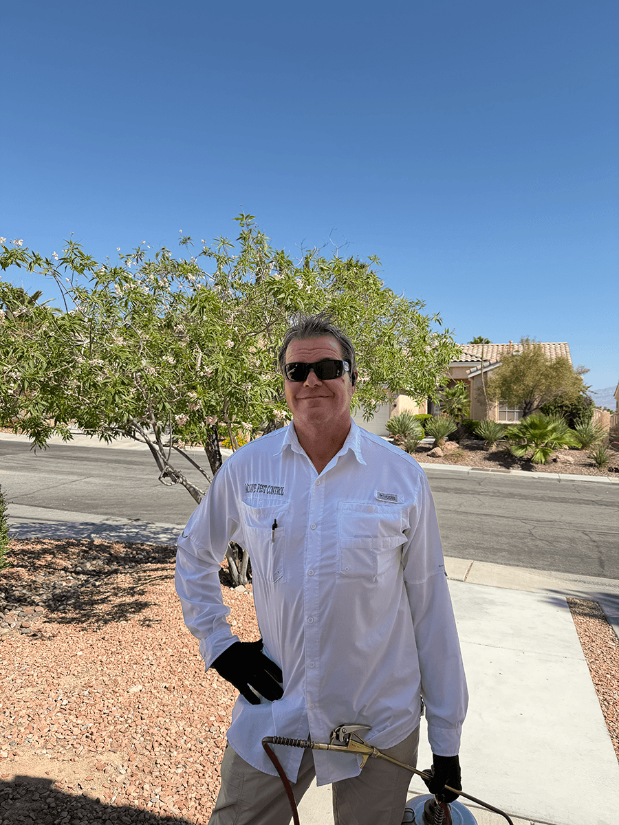 A man in sunglasses wearing a white button-up shirt standing outdoors on a sunny day, with desert plants and a blue sky in the background.