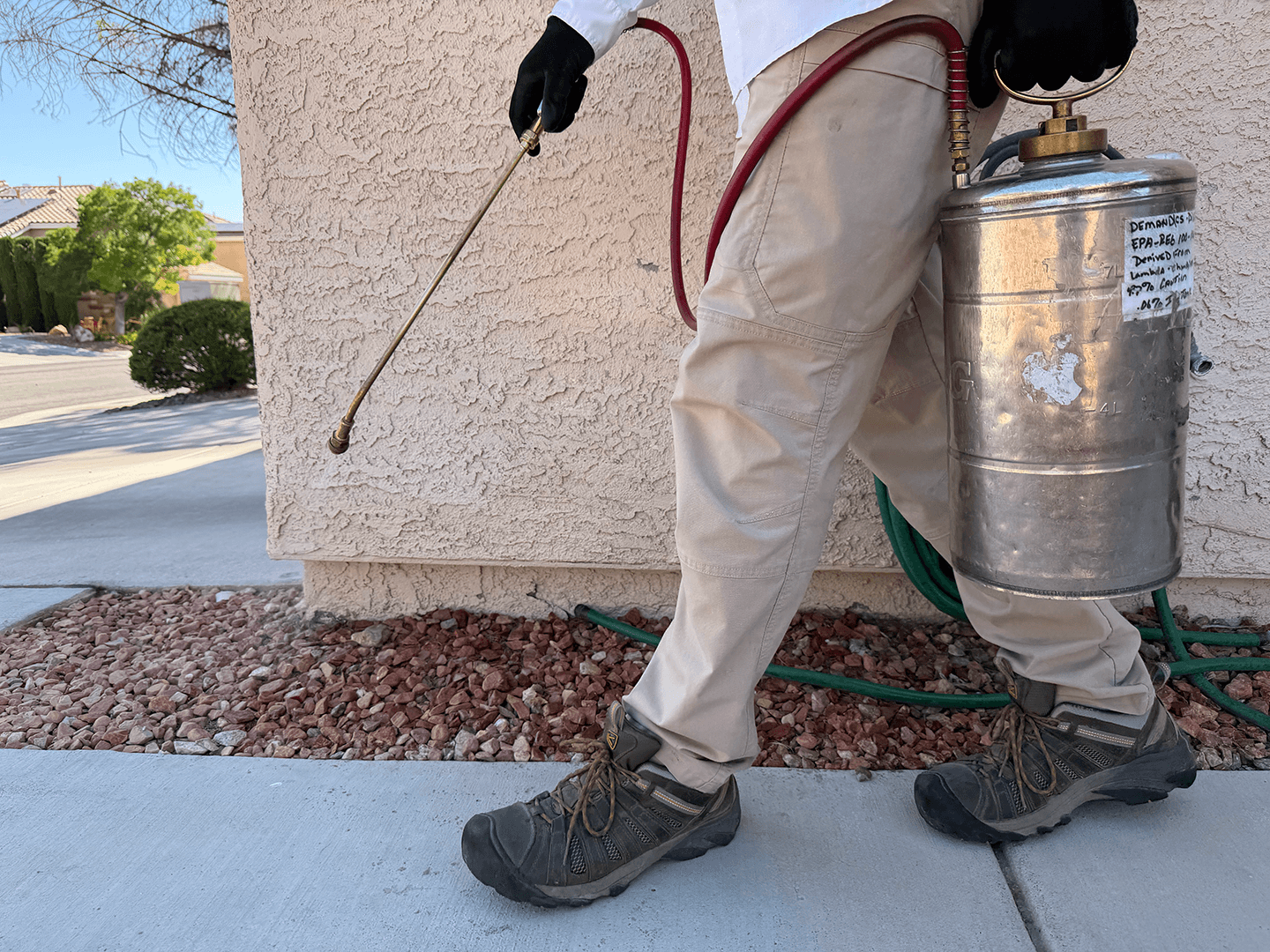 Close-up of a person wearing beige work pants and black safety shoes, holding a spray nozzle connected to a metal container, standing near a textured beige wall on a sidewalk with small rocks and a green garden hose.