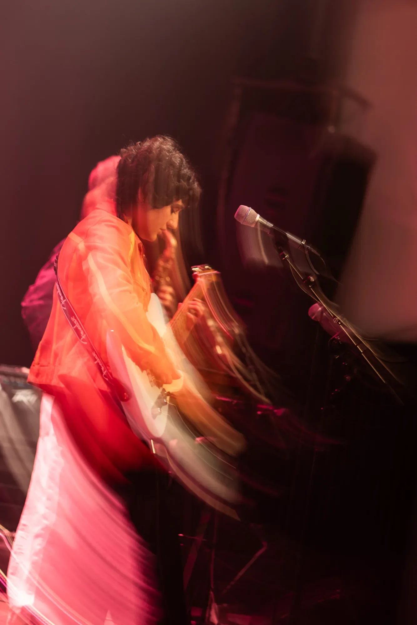 Camille Santacroce playing an electric guitar on stage, with a microphone nearby, motion blur creates a sense of movement.
