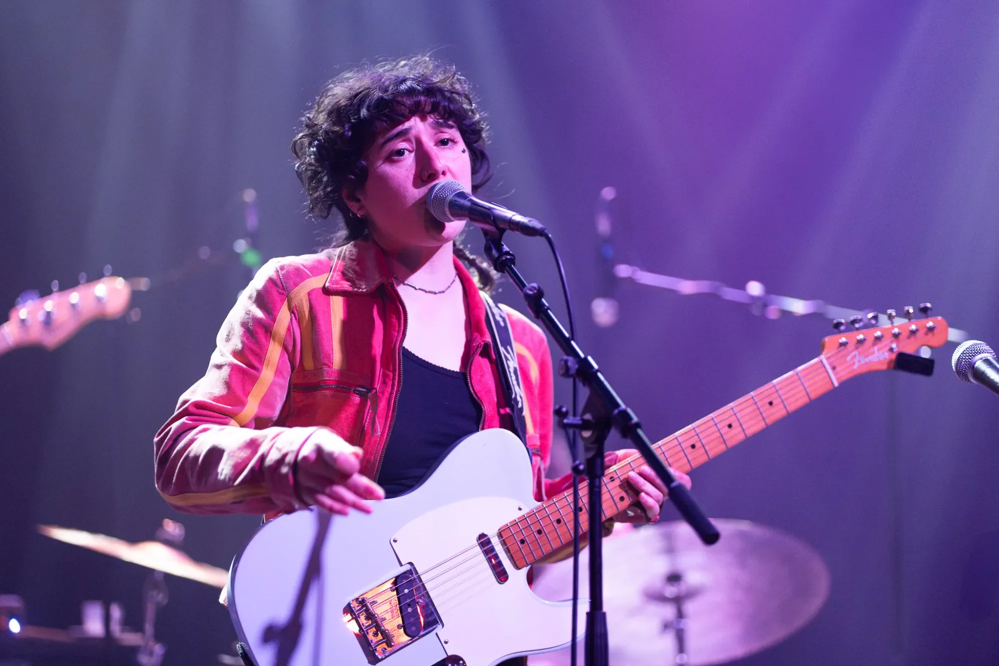 Singer-songwriter Camille Santacroce singing into a microphone while playing a white electric guitar on stage, with purple stage lights in the background.
