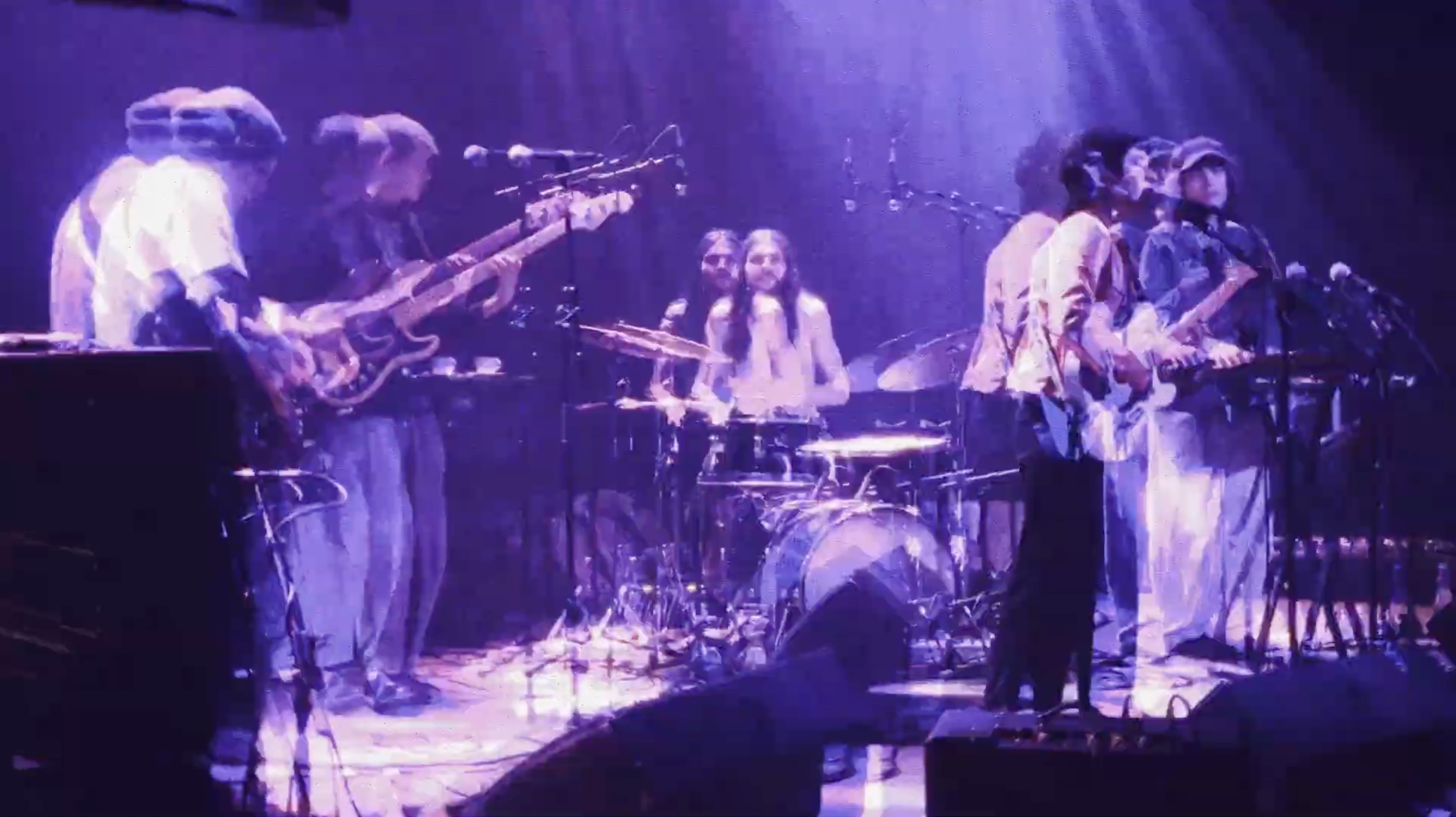 A live music performance with multiple musicians including Camille Santacroce playing instruments on stage, including guitars, drums, and keyboards, under purple stage lighting.