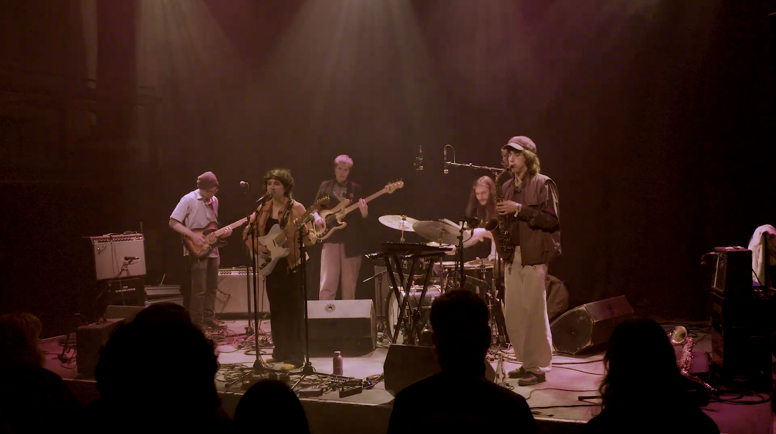 A band of six musicians performing on stage with various instruments including guitars, drums, and a saxophone, under dim lighting in a dark venue with an audience seated in front.