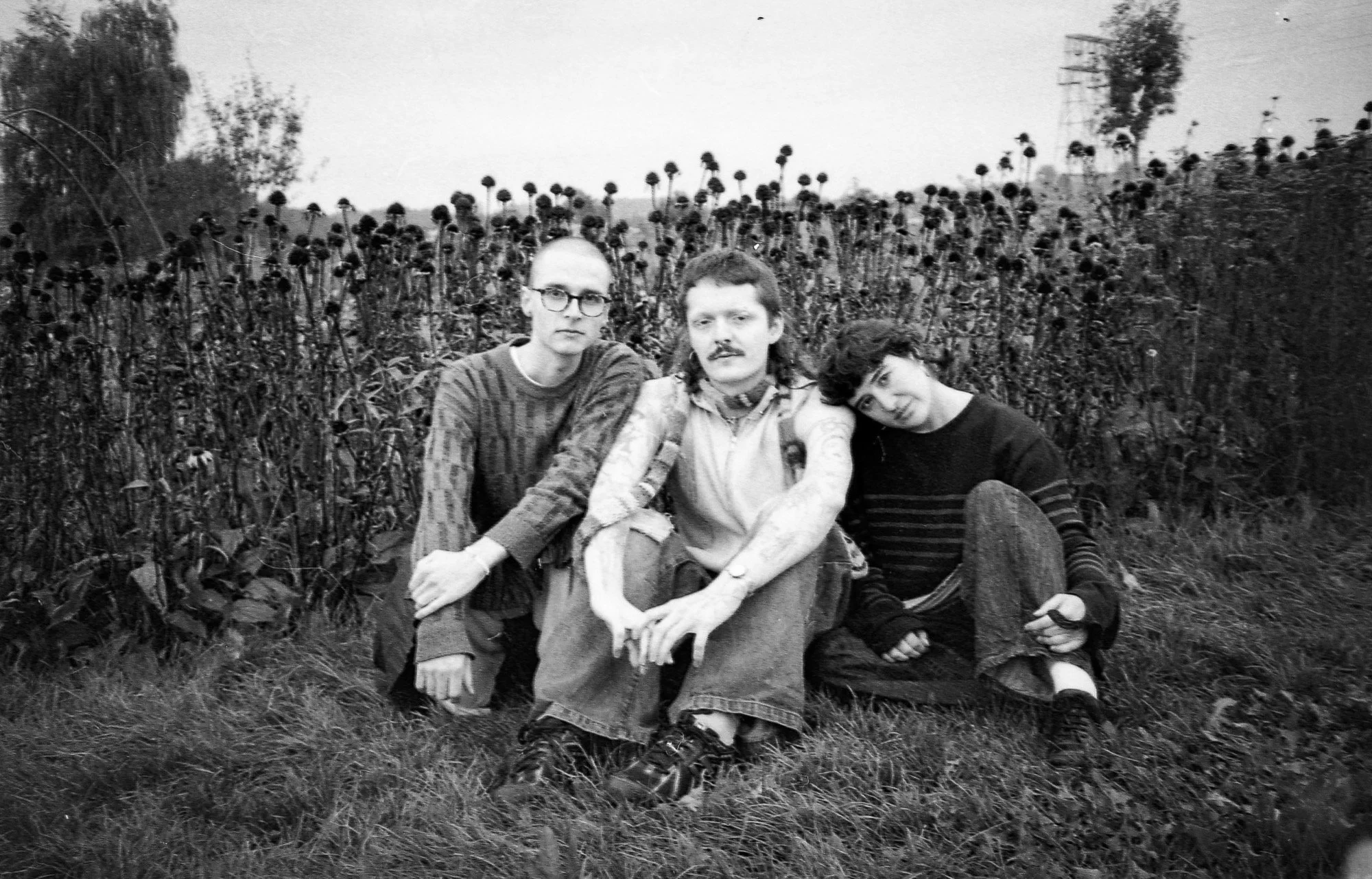 Band members of ottotto including Camille Santacroce. Black and white photo of three young people sitting on grass in front of a withered flower field.